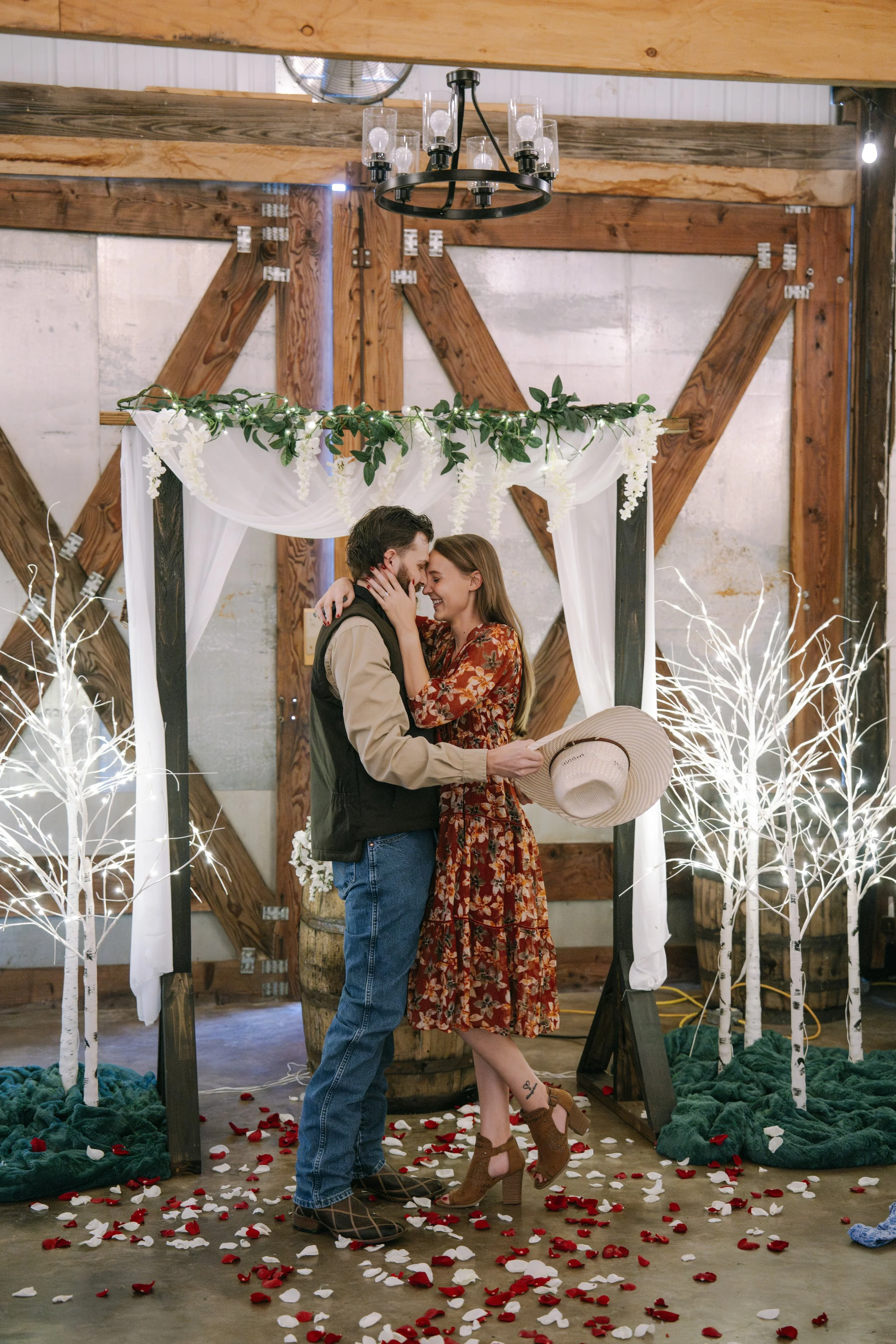 A couple with closed eyes in romantic embrace on a decorated wedding altar, with rose petals scattered on the floor, surrounded by white trees and rustic wooden beams.