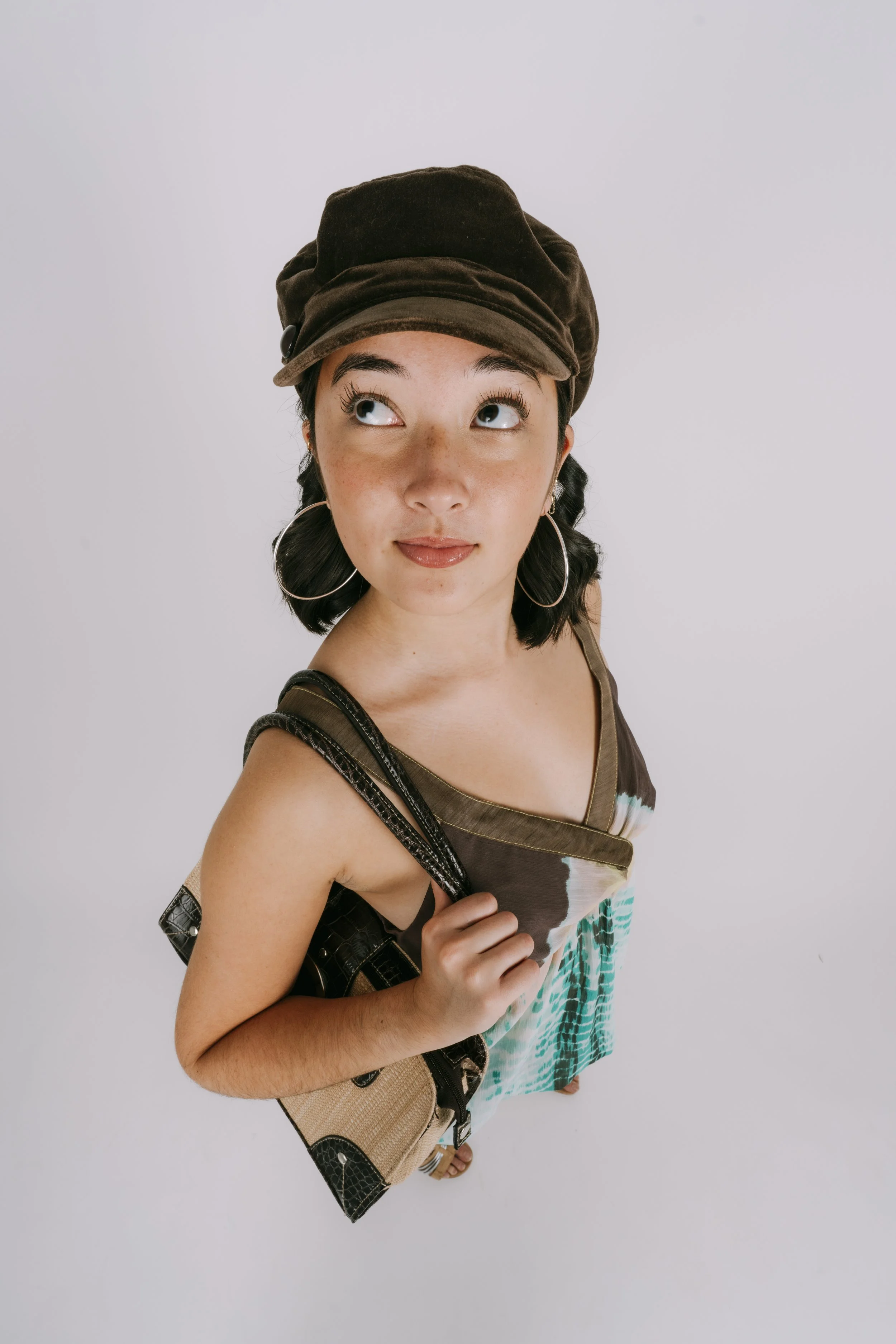 A young woman with dark hair, wearing a brown hat, large hoop earrings, a sleeveless dress, and sandals, standing against a plain white background, looking upward and slightly to the side.