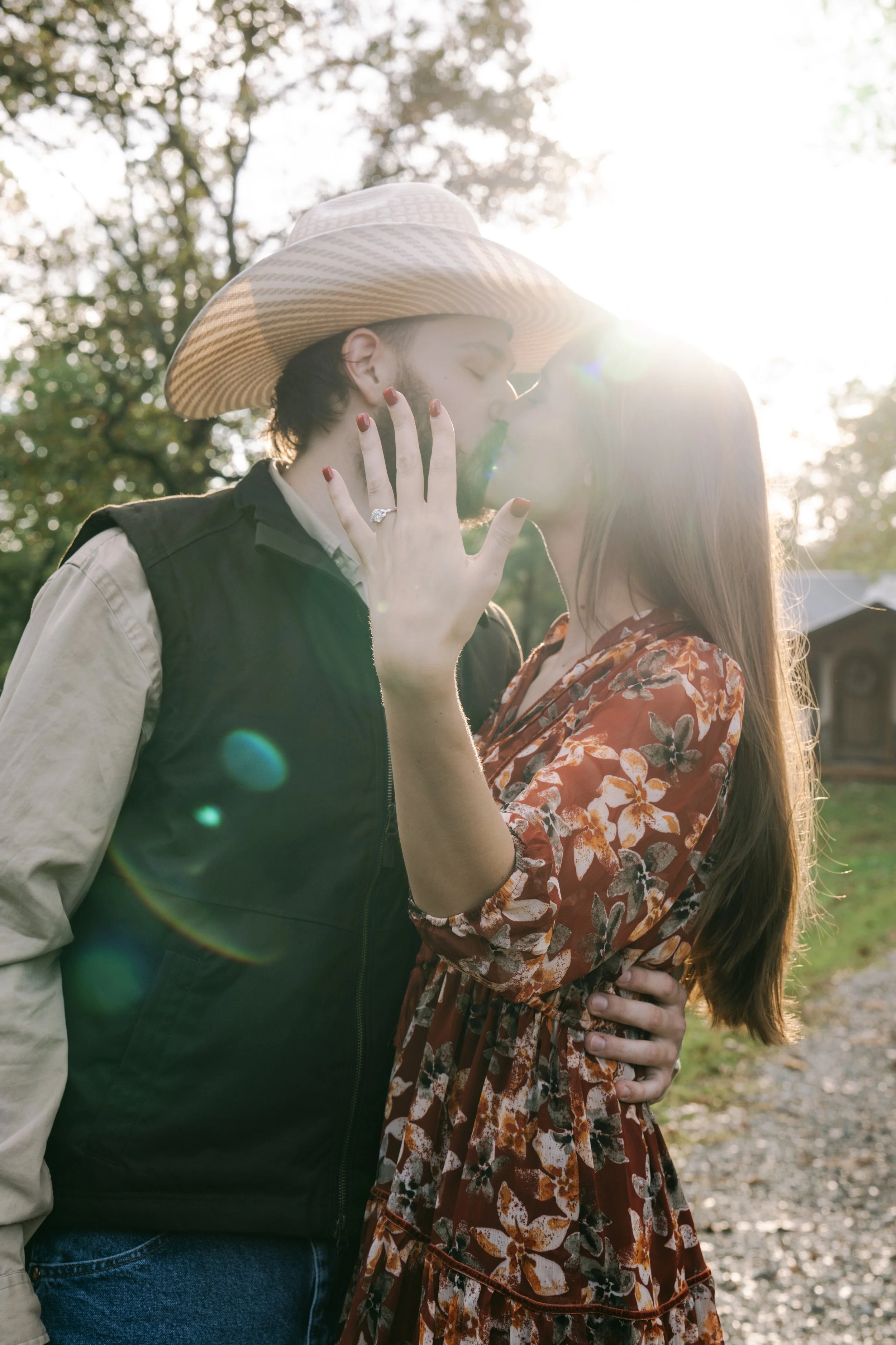 A couple kissing outdoors in the sunlight, with the man wearing a cowboy hat and a vest, and the woman in a floral dress, with her hand showing an engagement ring.