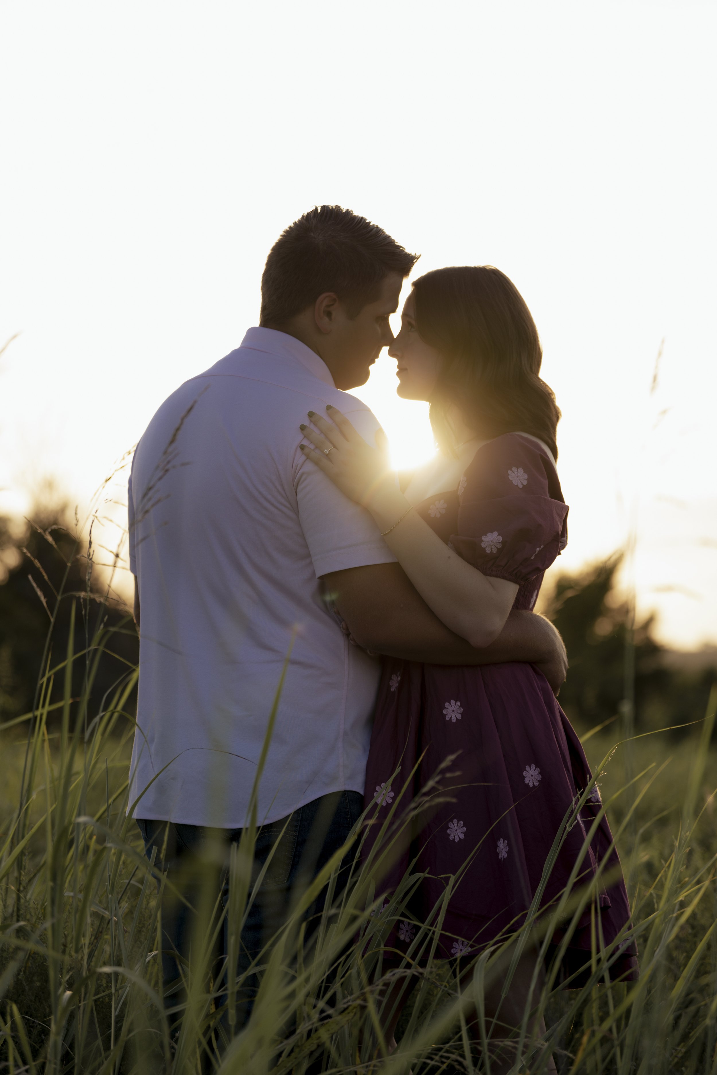 A couple embraces in a grassy field during sunset, faces close, backlit with warm sunlight.