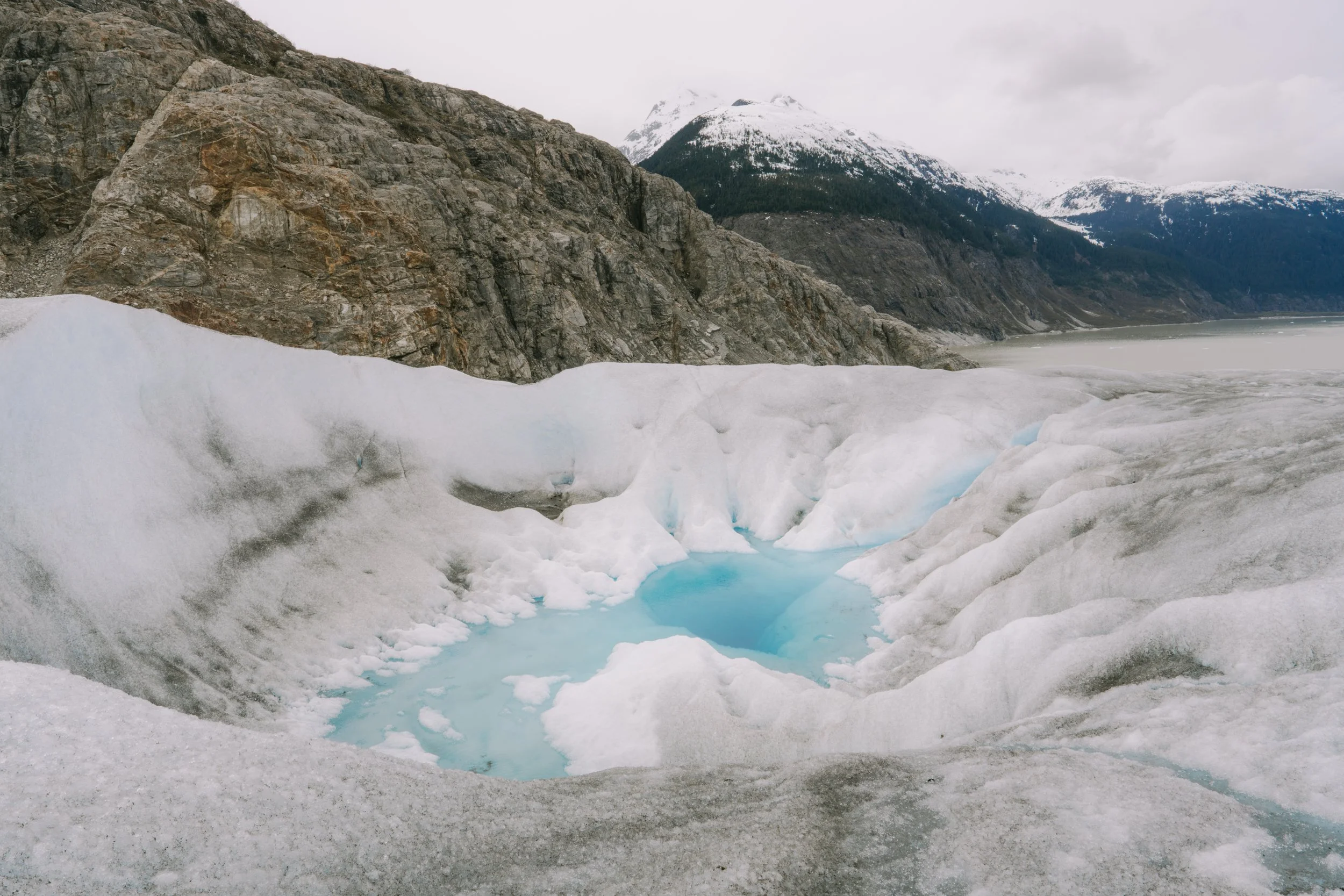 A glacier with melting ice and blue water in front of mountainous landscape with snow-capped peaks.