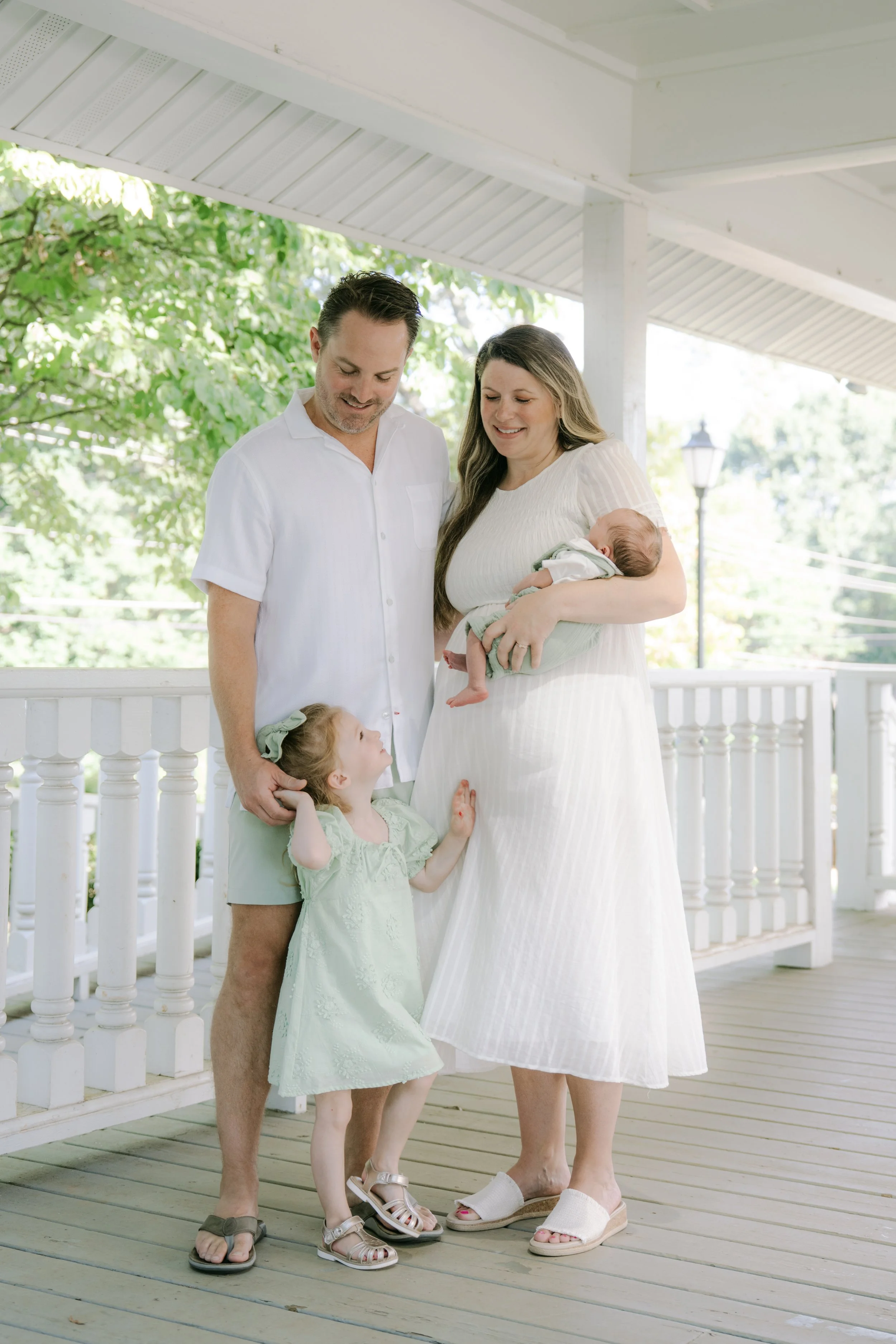Family portrait of a man, woman, and two young girls on a porch, with a baby. The woman is holding the baby, and the older girl is looking up at the woman while the man is holding the younger girl.