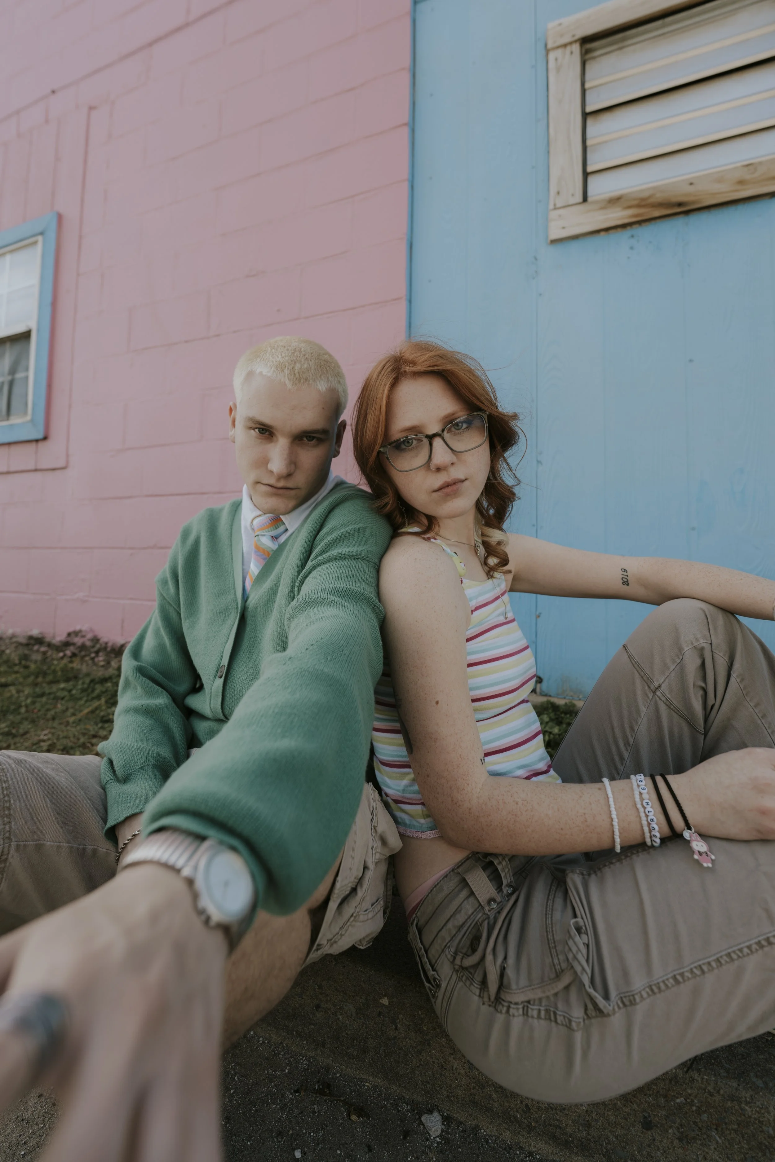 A young man and woman sitting on the ground outside, leaning against a pastel pink and blue wall with small windows. The man has short, bleach-blond hair, is wearing a green cardigan, white shirt, and striped tie, and is taking a selfie. The woman ha