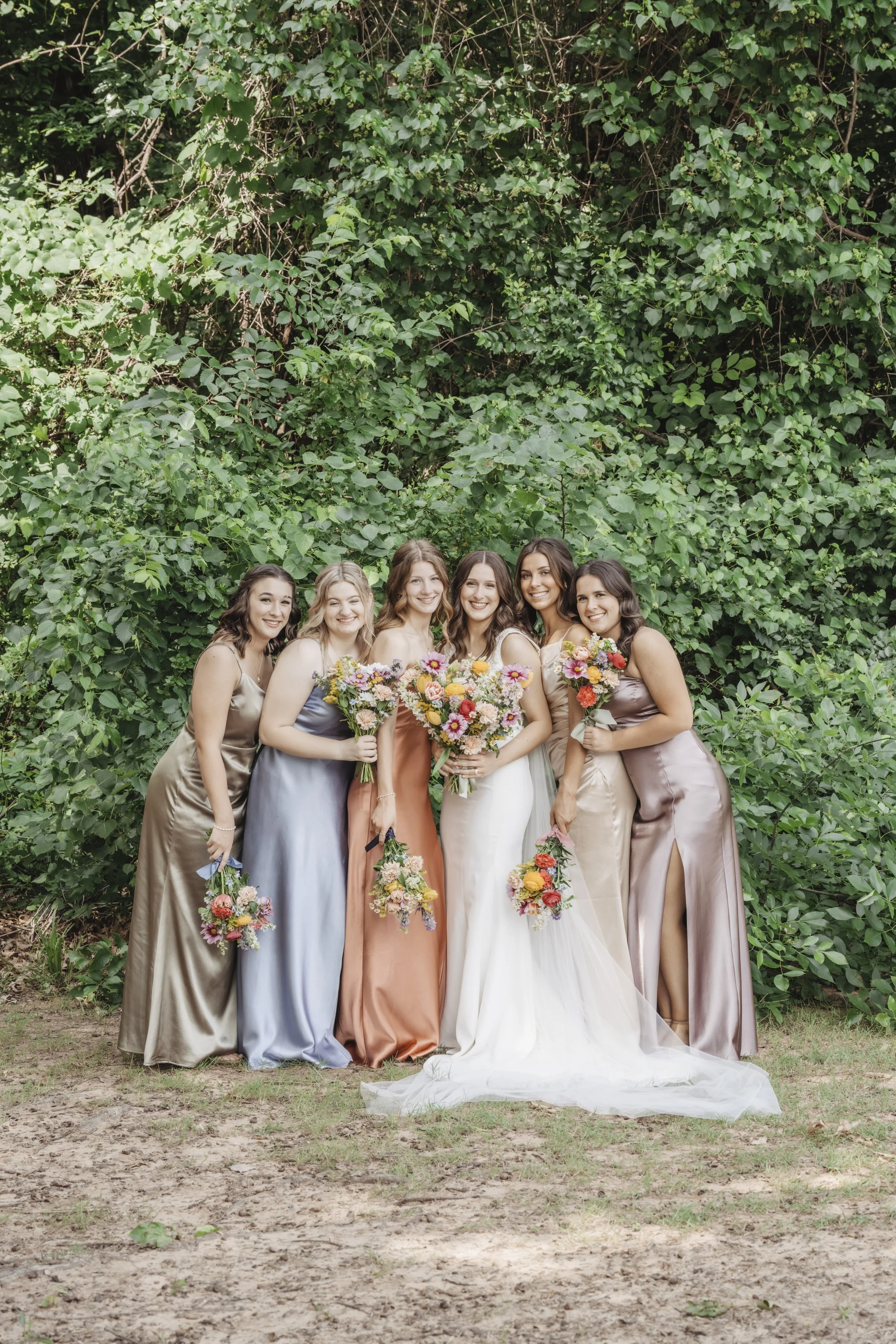 Group of six women in colorful dresses holding bouquets of flowers, standing outdoors in front of lush green foliage.