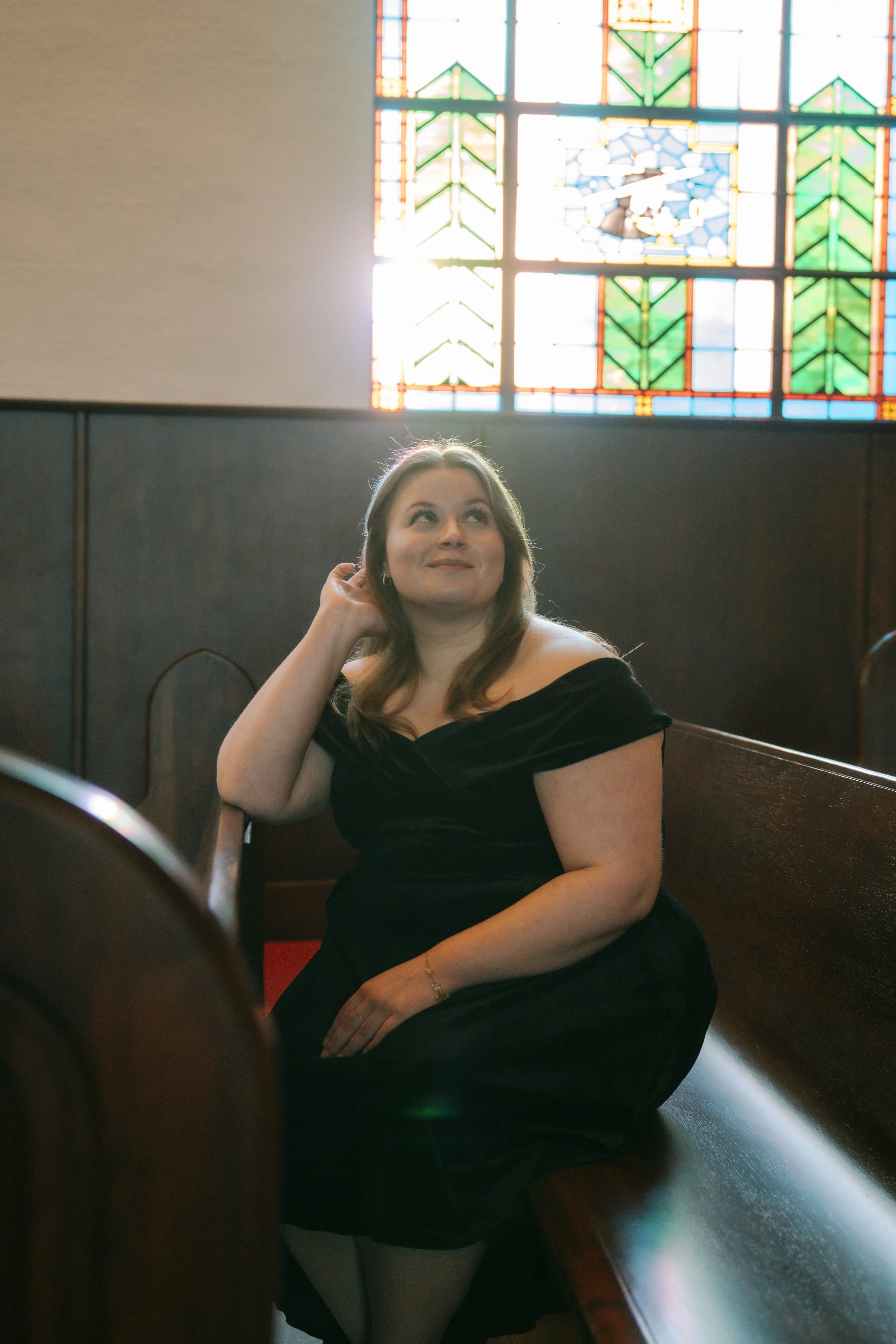 Woman in a black off-the-shoulder dress sitting on a church pew inside a church with stained glass windows.