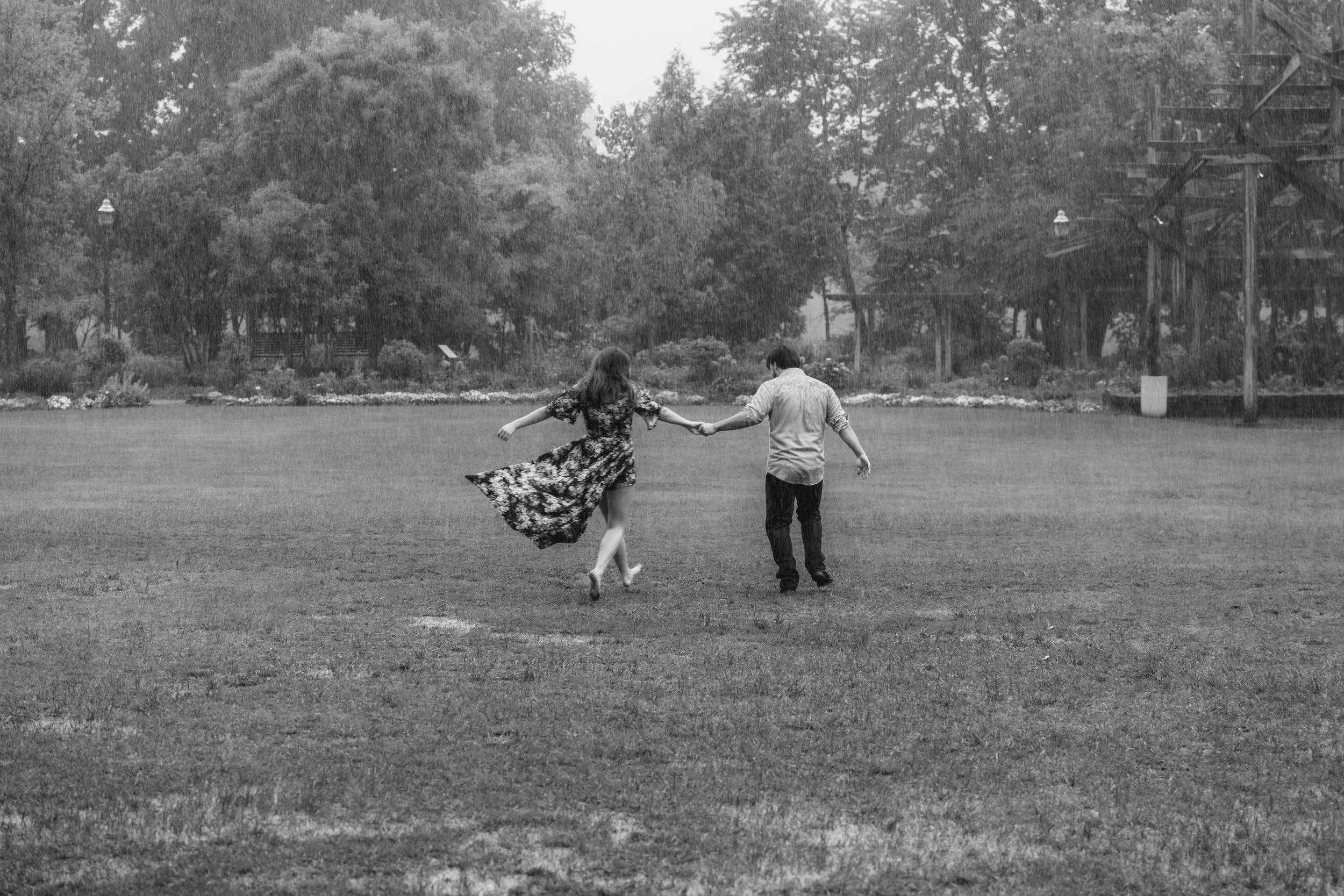 A black and white photo of a young couple holding hands, walking and running on a grassy field in a rainstorm, surrounded by trees.