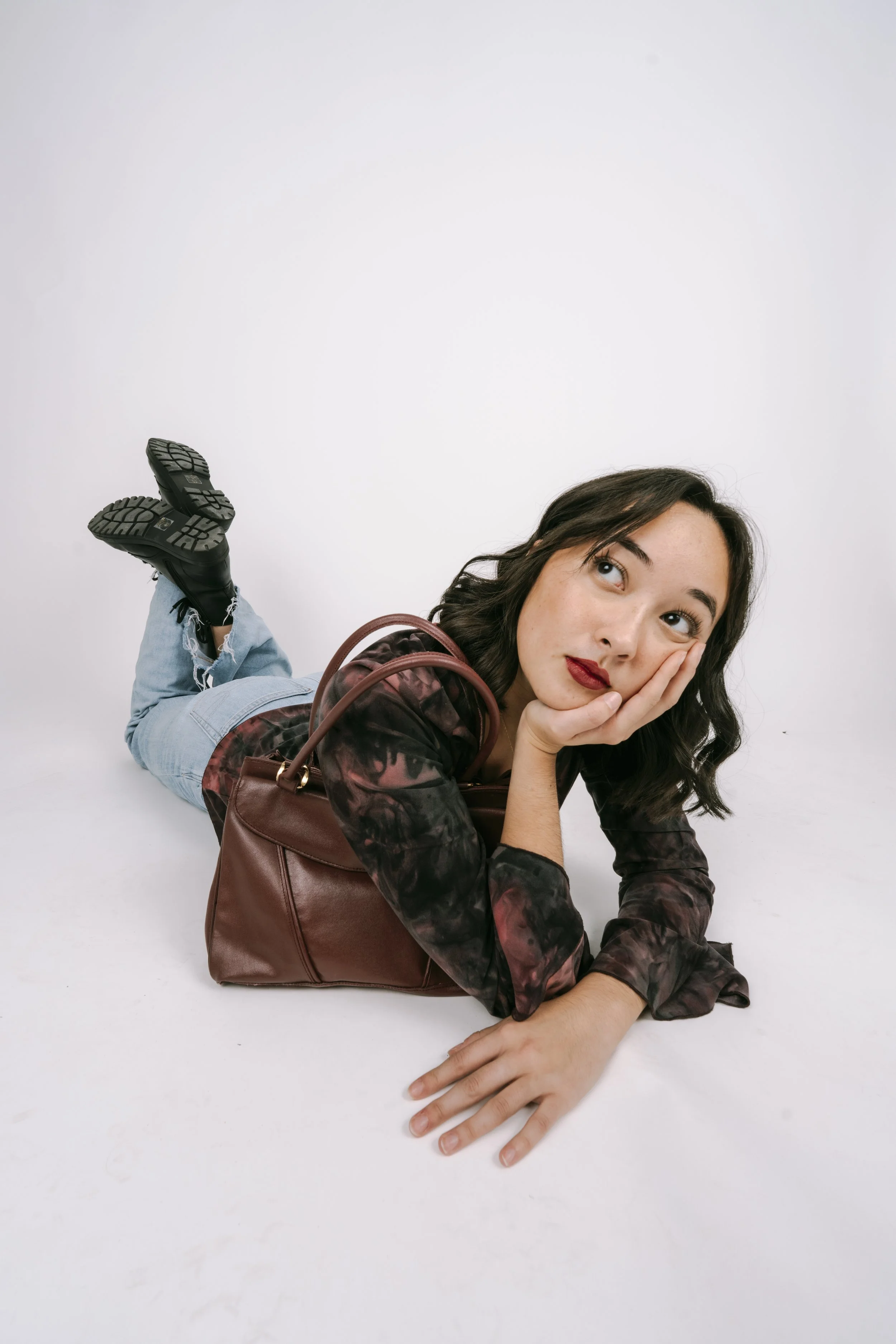 Young woman lying on her stomach on a white background, resting her face on her hand, with a brown handbag nearby.