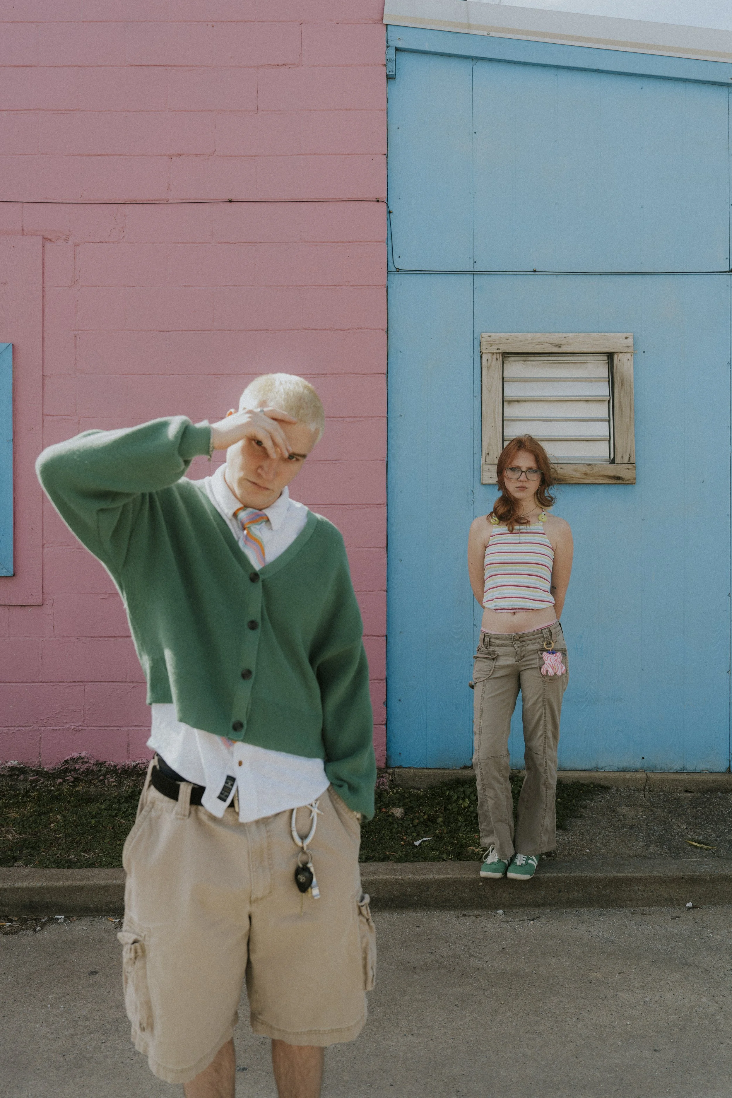 A young man with bleached blonde hair and a young woman with red hair and glasses standing in front of a colorful pink and blue building.