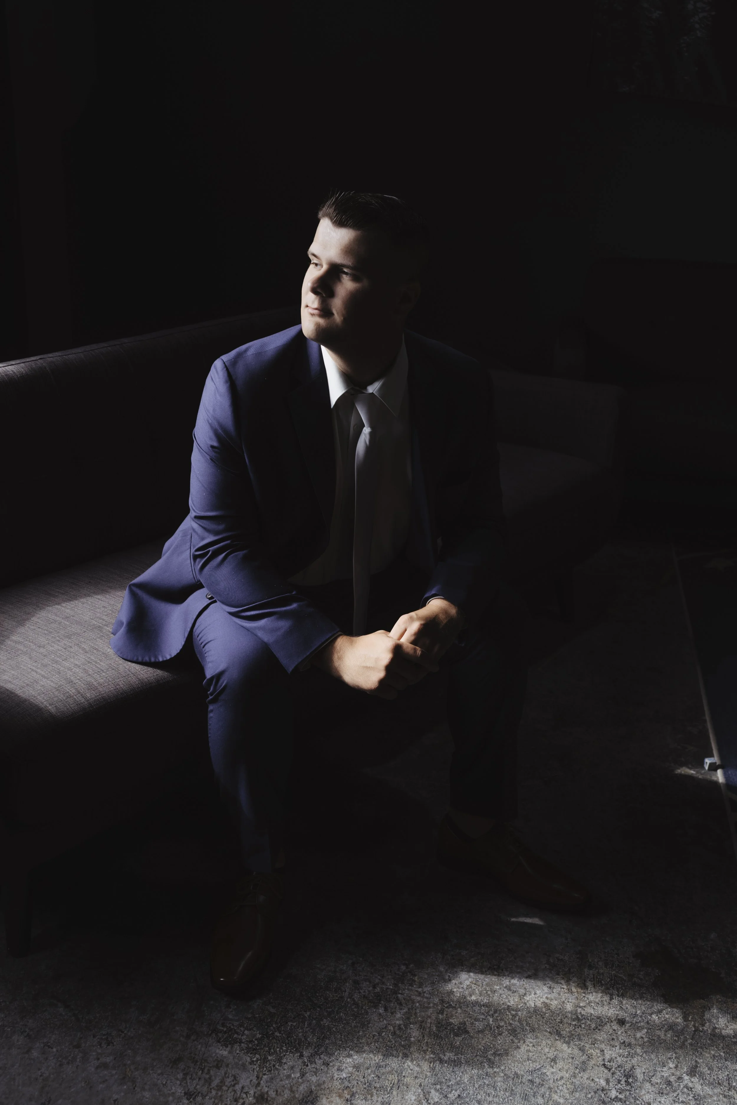 A man in a dark blue suit sitting on a dark couch, looking to the side with light hitting his face, in a dimly lit room.