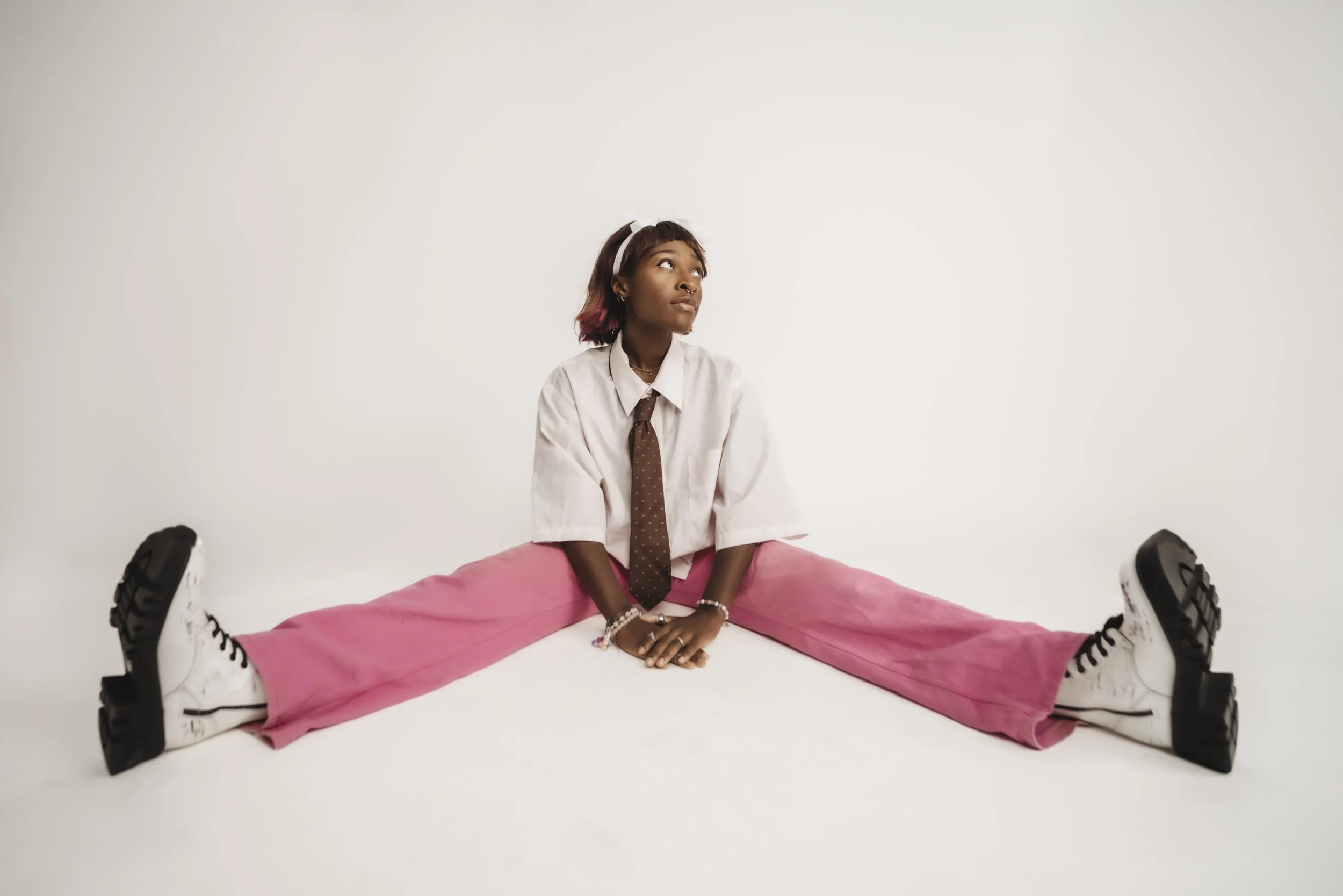 Young woman sitting on the floor with legs spread apart, wearing white sneakers, pink pants, a white shirt, and a brown tie, looking up and to the side against a plain white background.