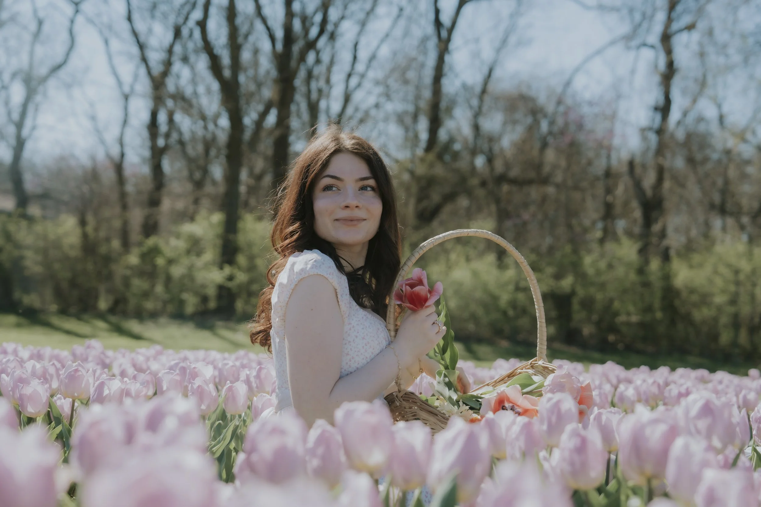 A woman with brown hair and a light dress holds a pink tulip while sitting in a field of pink tulips with trees in the background.