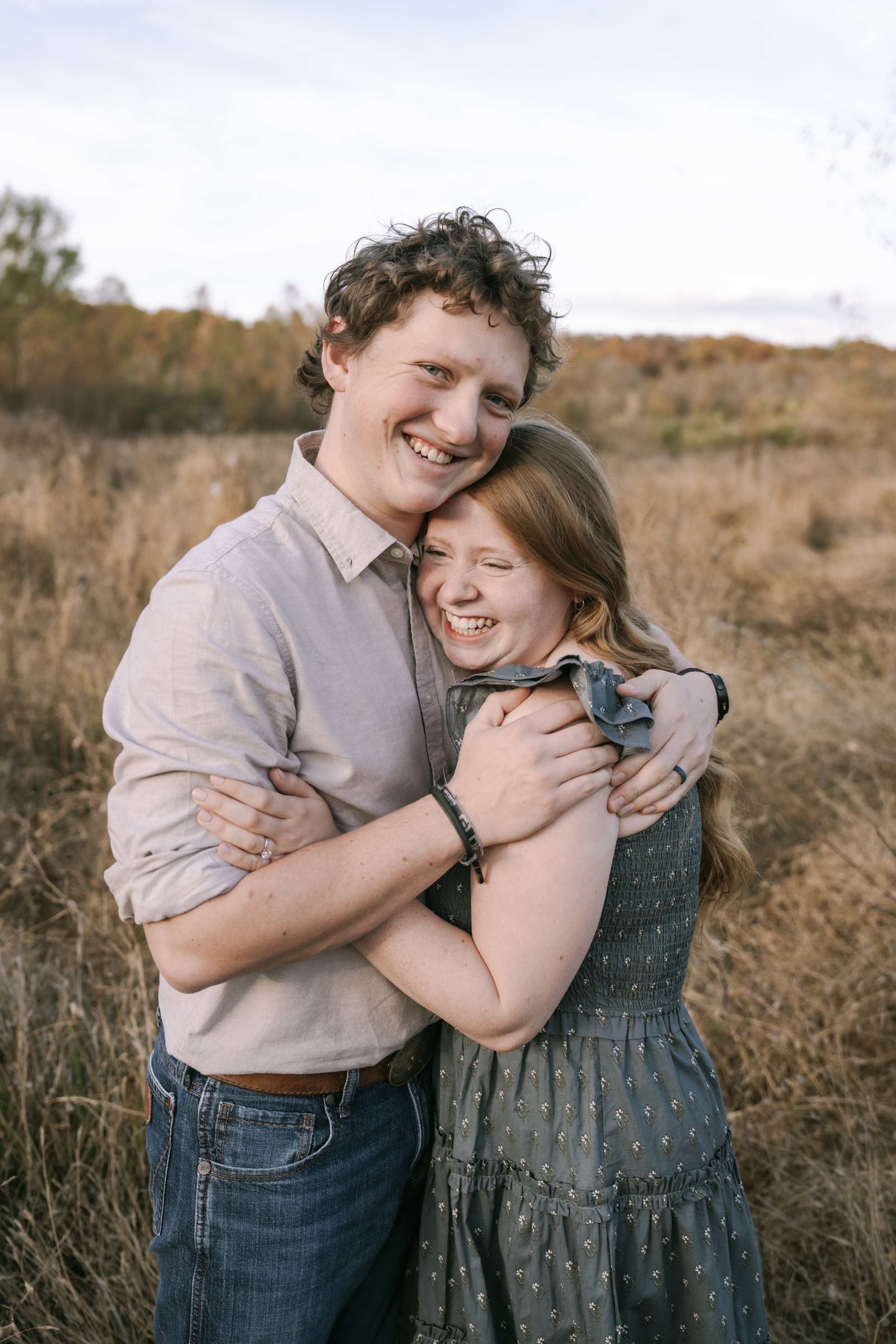 A young man and woman are happily hugging outdoors in a field with trees and a cloudy sky in the background.