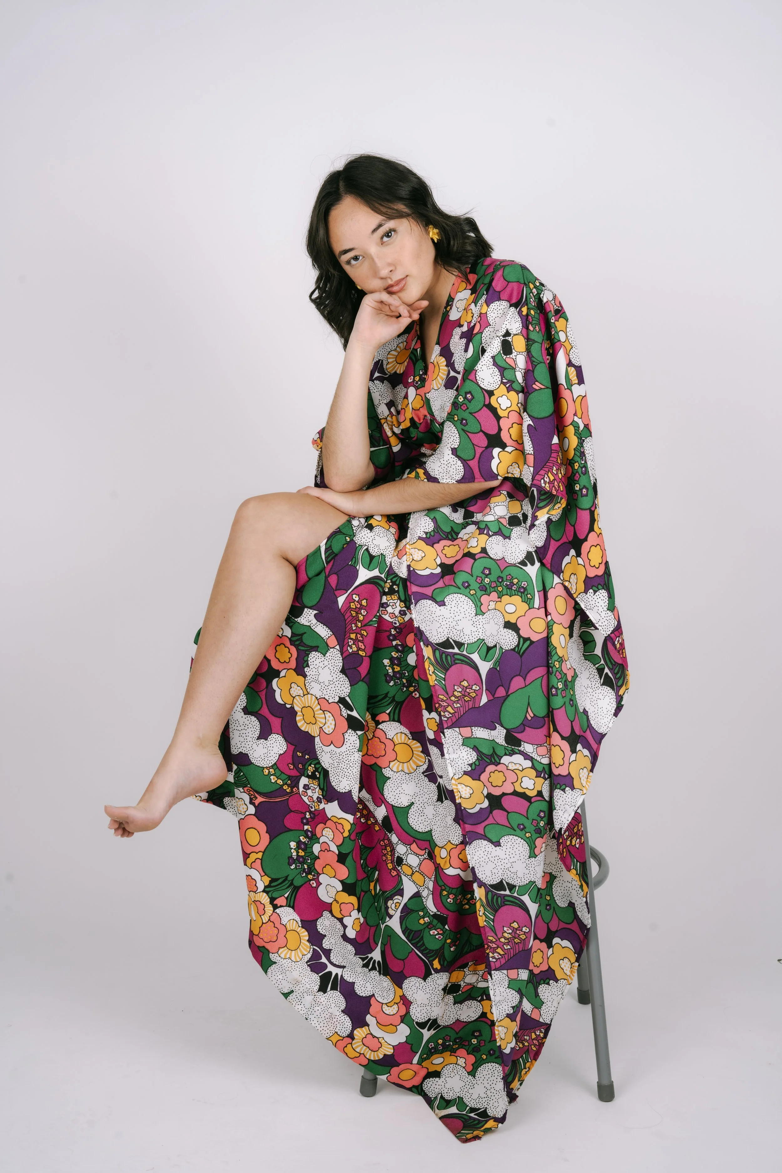 Woman sitting on a stool with her left leg crossed over her right, wearing a colorful floral kimono-style garment, with a thoughtful expression, against a plain white backdrop.