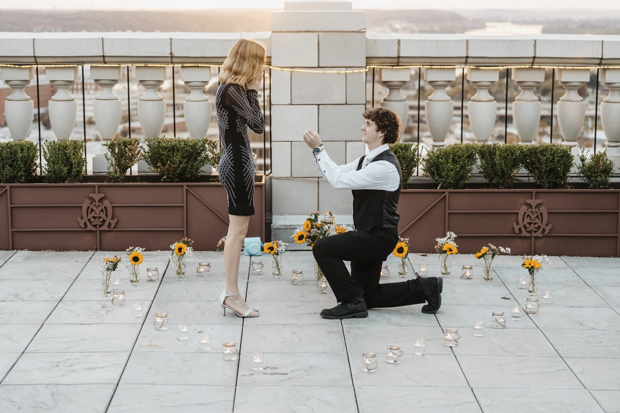 A man proposing to a woman on a rooftop patio during sunset, surrounded by flowers and candles.