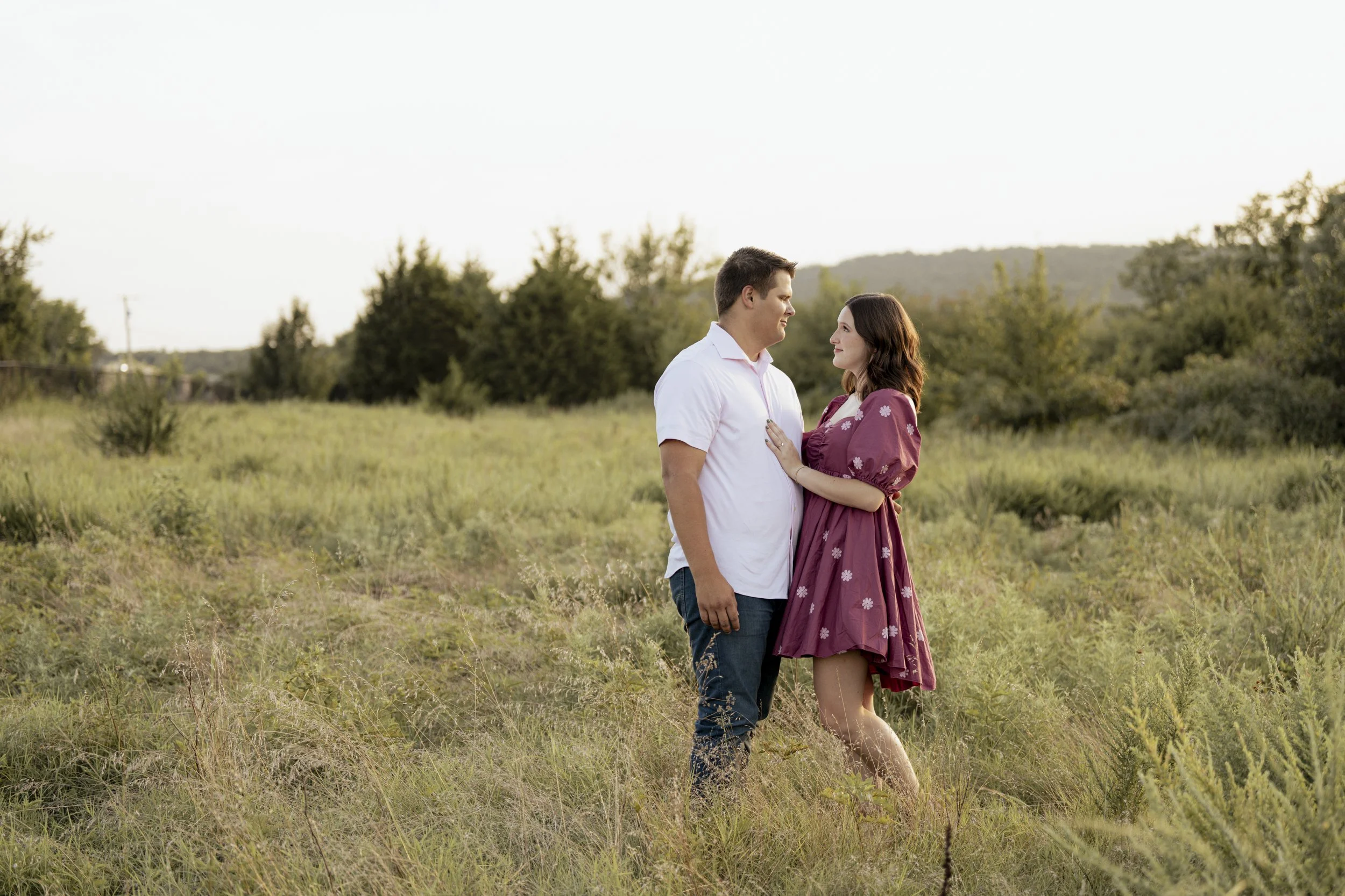 A couple standing close together in a grassy field, gazing into each other's eyes during sunset.