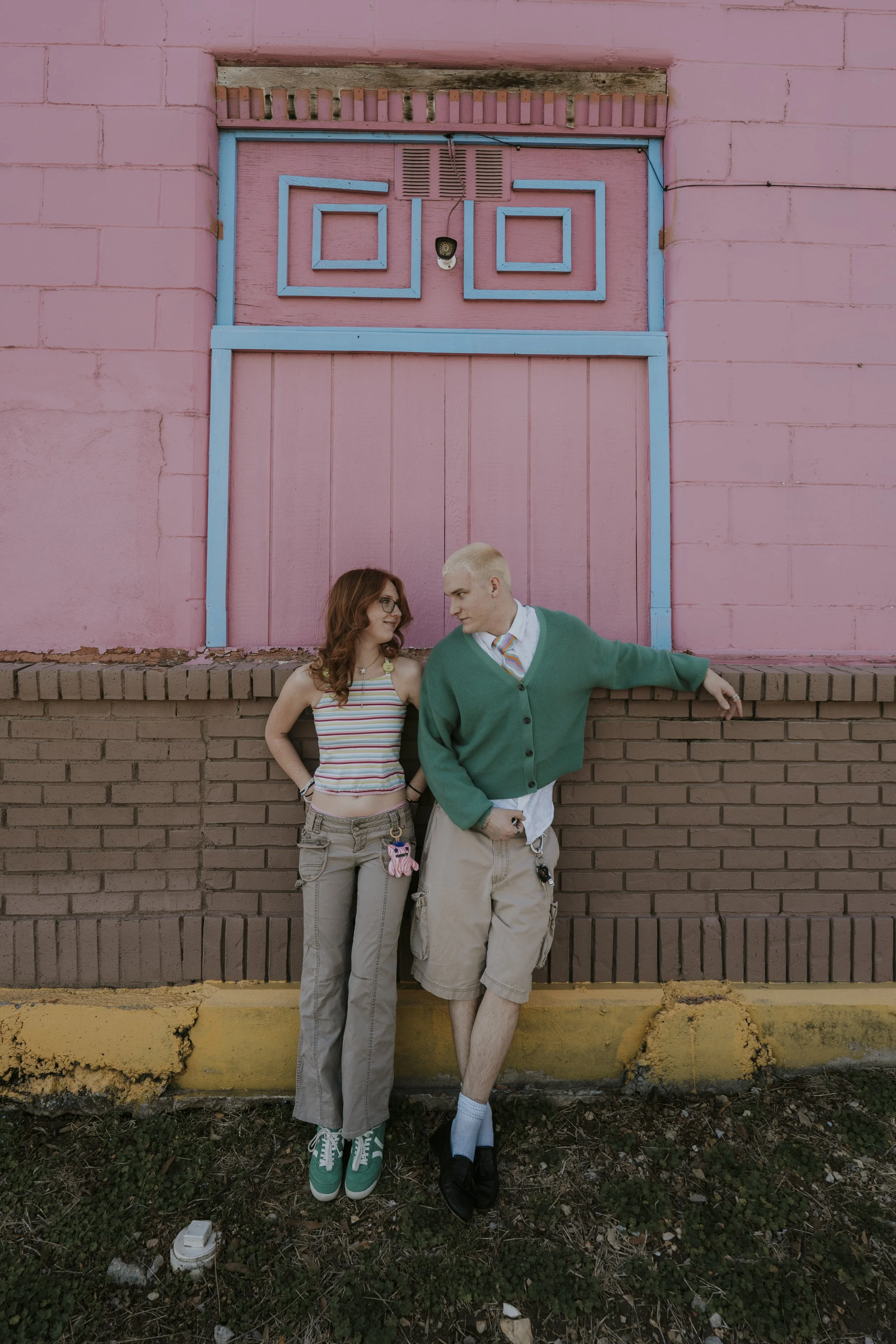 Two young adults, a woman with red hair and glasses wearing a striped tank top and beige cargo pants, and a man with bleached hair wearing a green cardigan, tie, and beige shorts, standing in front of a pink and brown building with a brick and painte