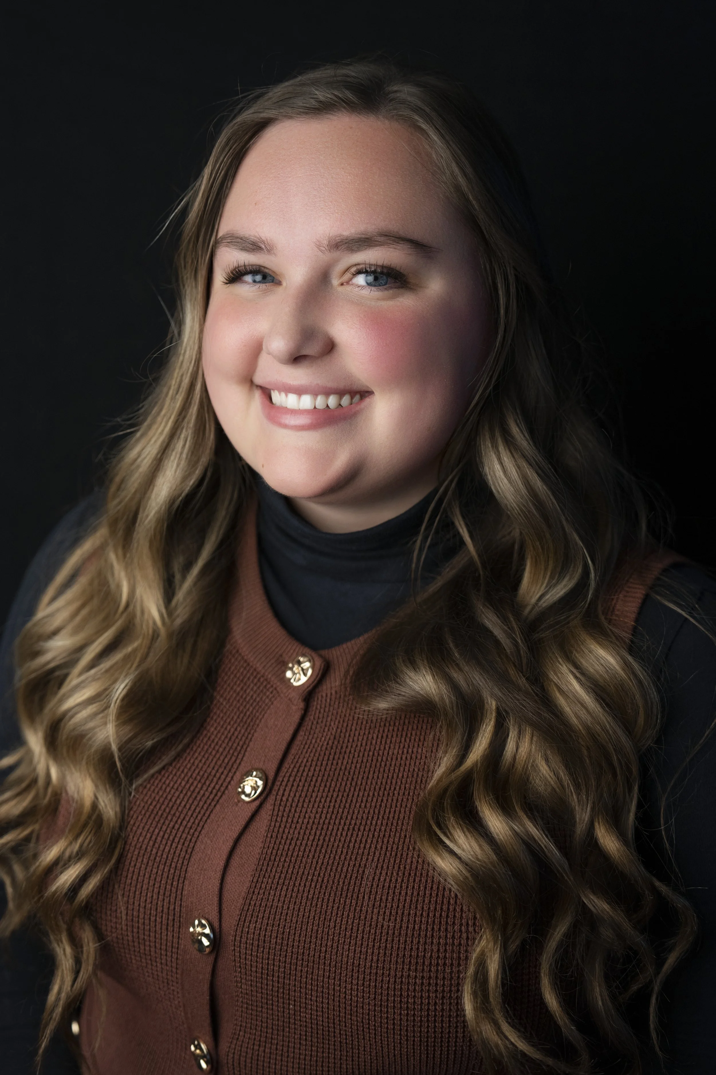 Portrait of a smiling young woman with long, wavy blonde hair, wearing a black turtleneck and a brown cardigan with gold buttons, against a dark background.