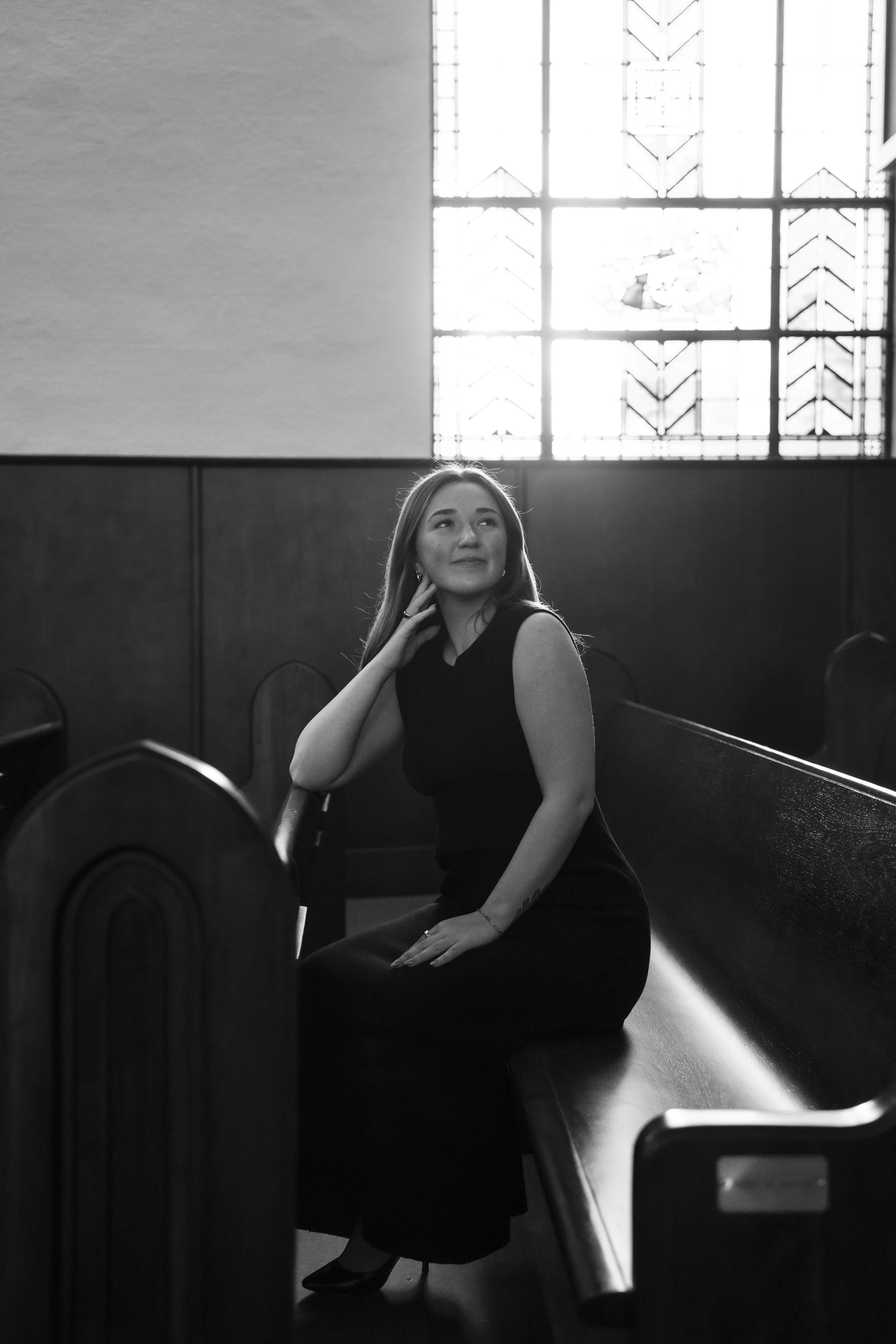 A young woman in a long, sleeveless black dress sitting on a church pew inside a dimly lit church with a large stained glass window behind her.