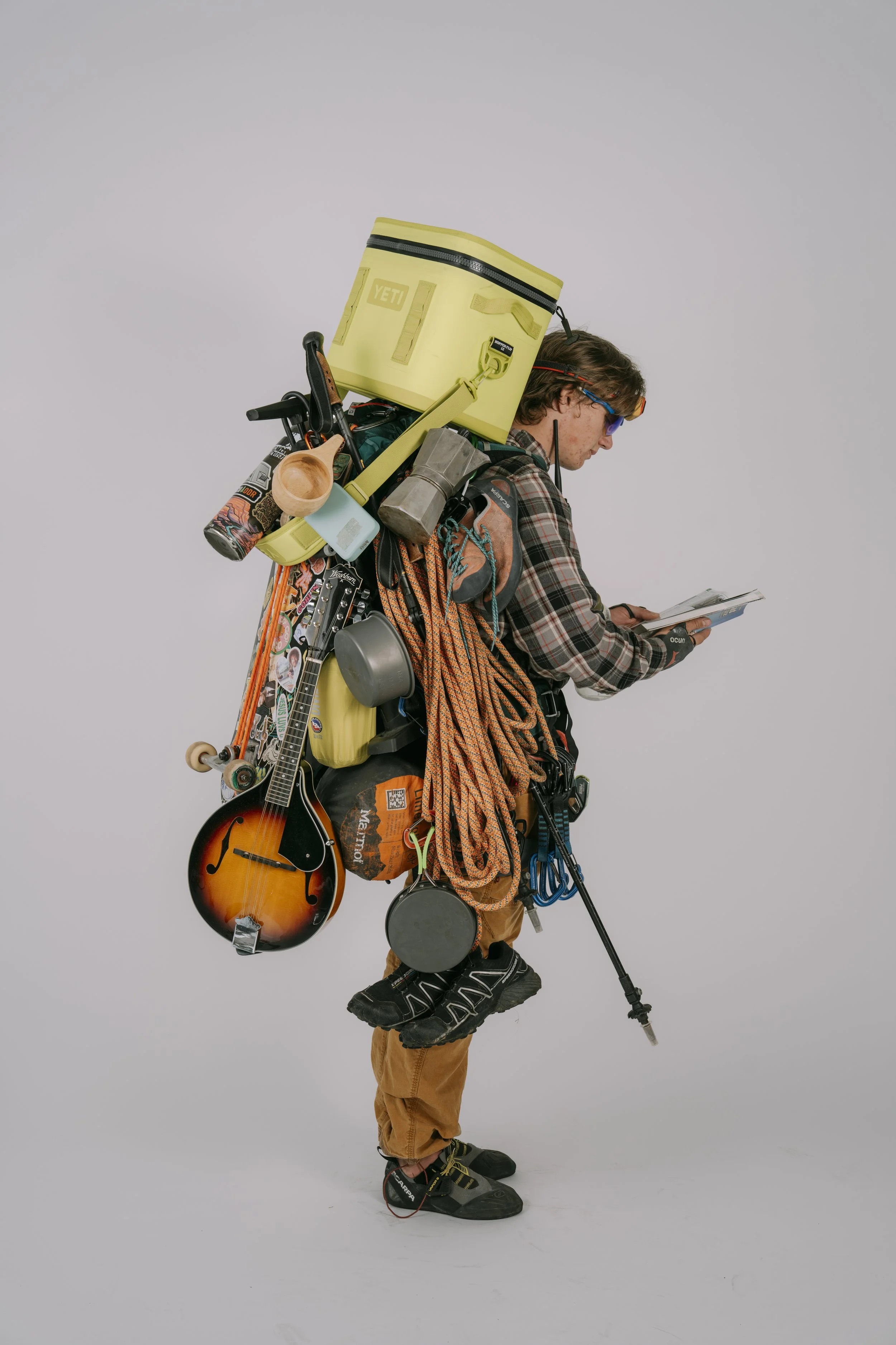 A man with a large backpack filled with camping and climbing gear, holding a booklet or map, standing against a plain gray background.