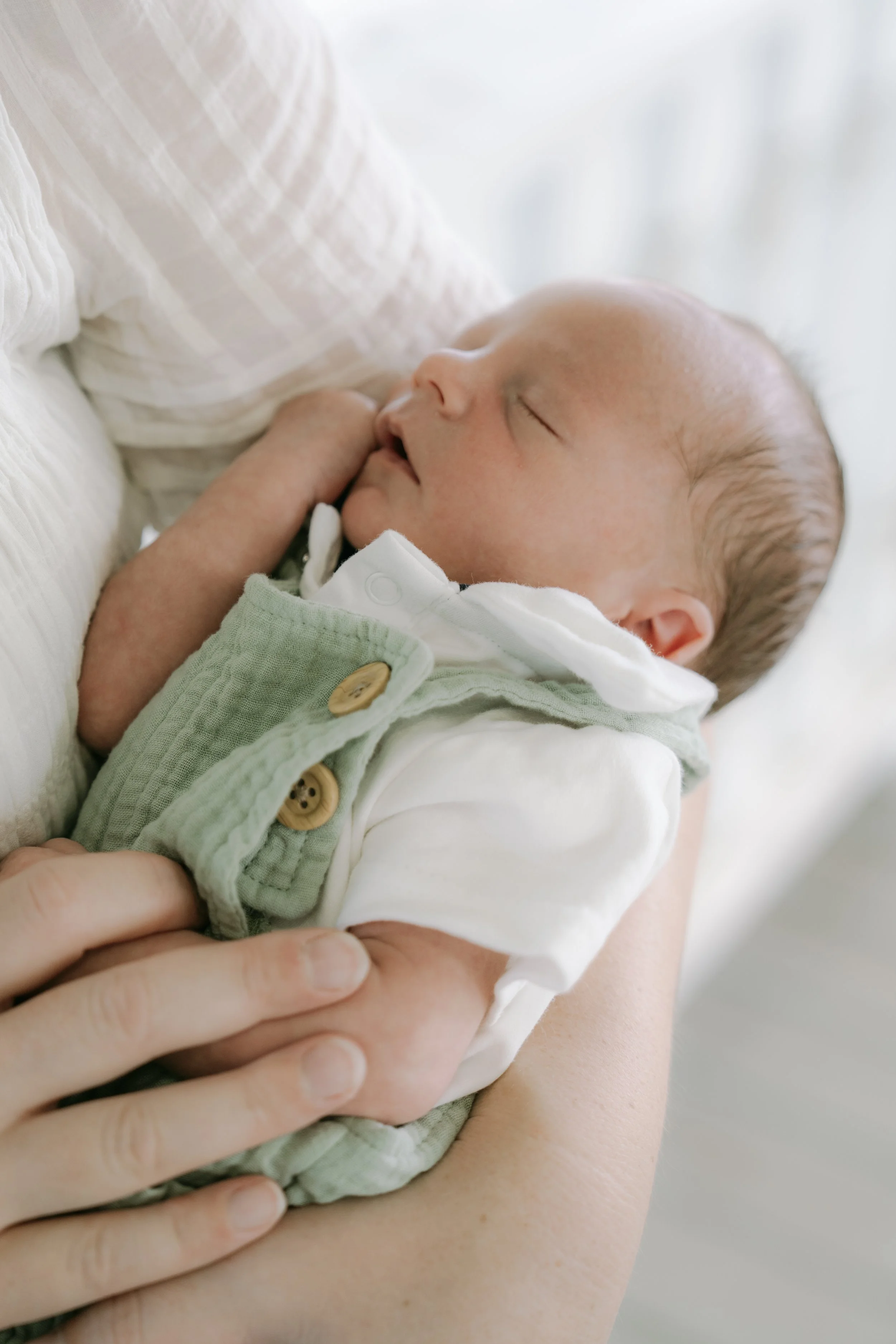 A sleeping baby being gently held by an adult, wearing a white shirt and light green vest with wooden buttons, close-up of the baby's face and surroundings.