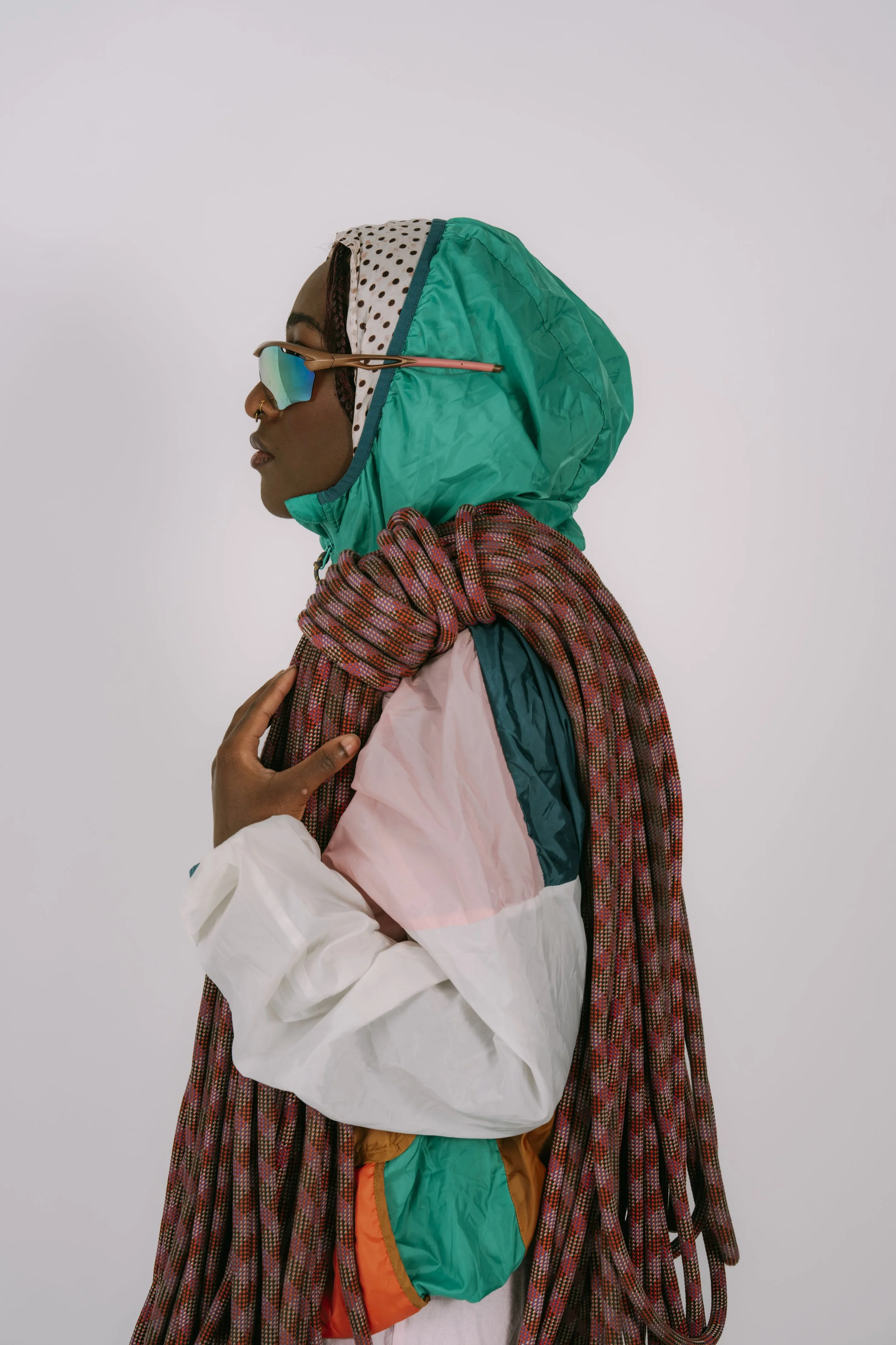 A woman wearing colorful, retro-style outdoor clothing, including a teal jacket with a hood, polka dot headband, mirrored sunglasses, and holding multiple climbing ropes over her shoulder.