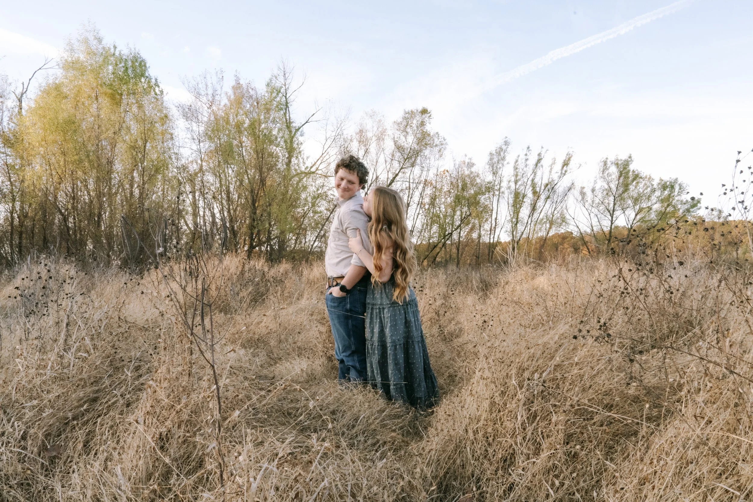 A young couple standing close together in a field of dried grass, with trees with fall foliage in the background, under a clear blue sky.