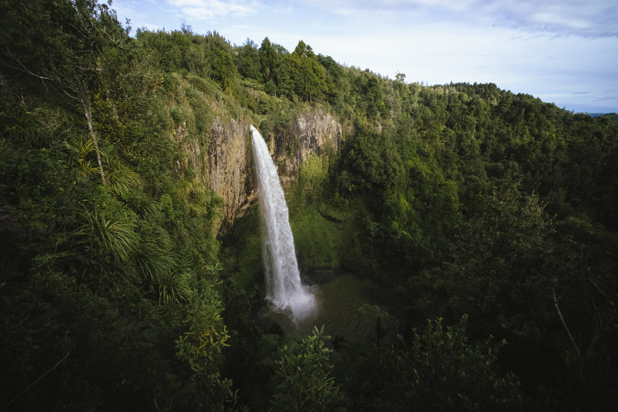 A tall waterfall cascading down a rocky cliff surrounded by lush green forest with a cloudy sky overhead.