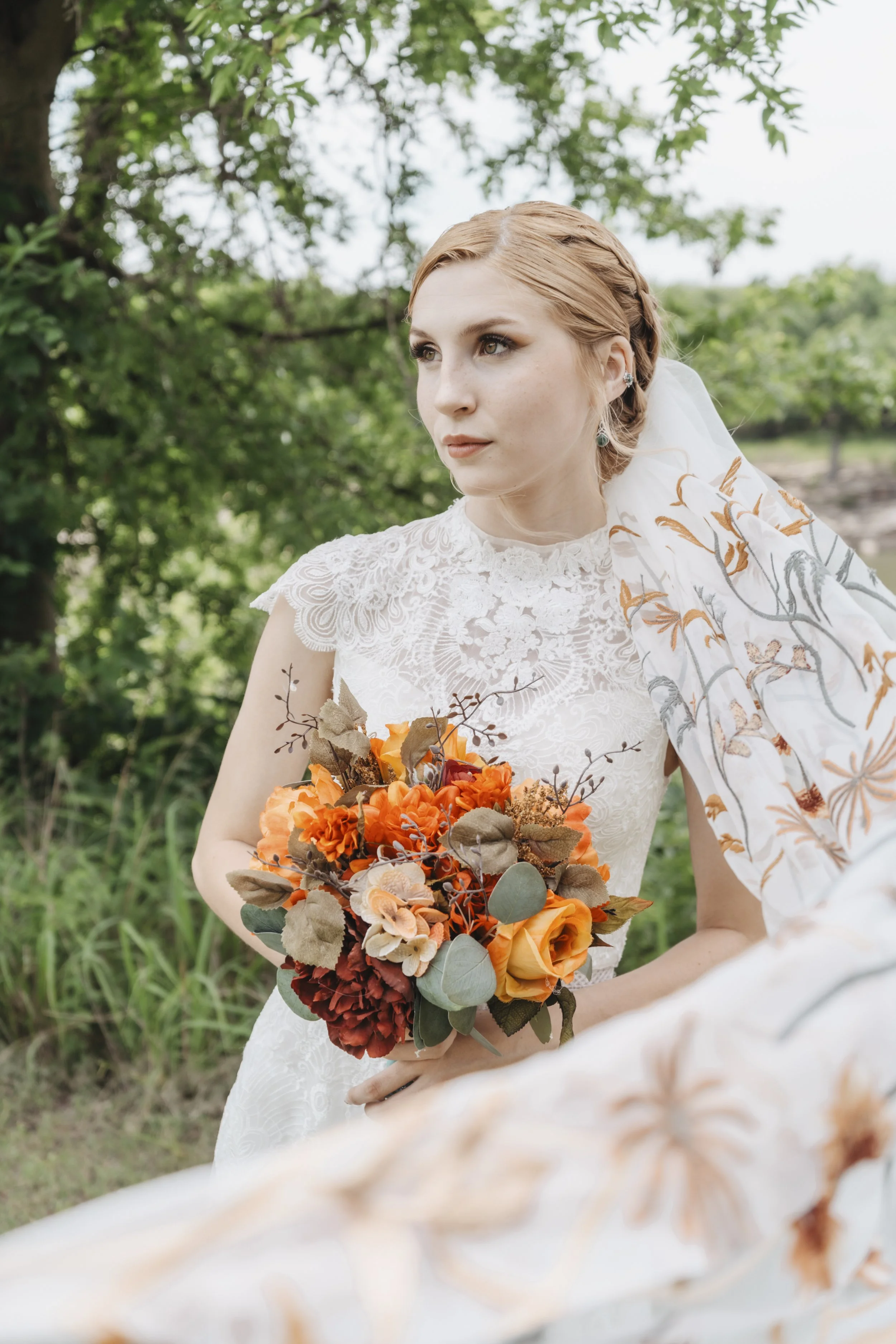 A bride with red hair, wearing a white lace wedding dress, holding a bouquet of orange, yellow, and red flowers with greenery, standing outdoors among green trees.
