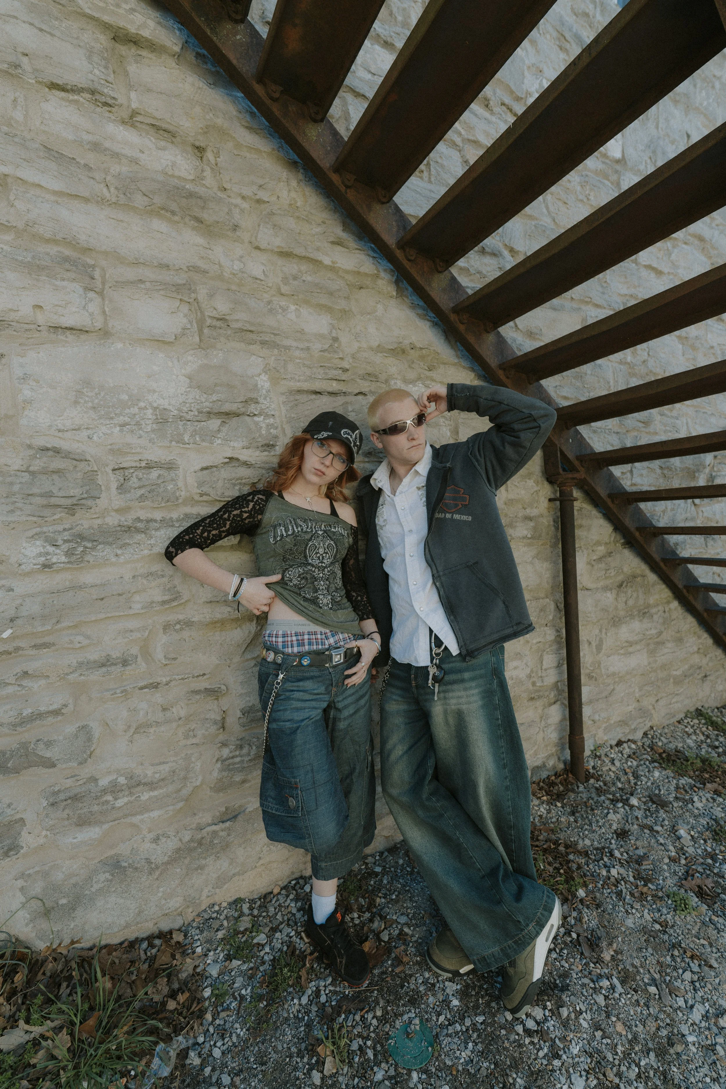 Two young people, a woman with red hair and a man with blond hair, standing against a stone wall near a rusty staircase. The woman wears glasses, a bandana, a black lace top, and baggy shorts with a plaid shirt layered underneath. The man wears sungl