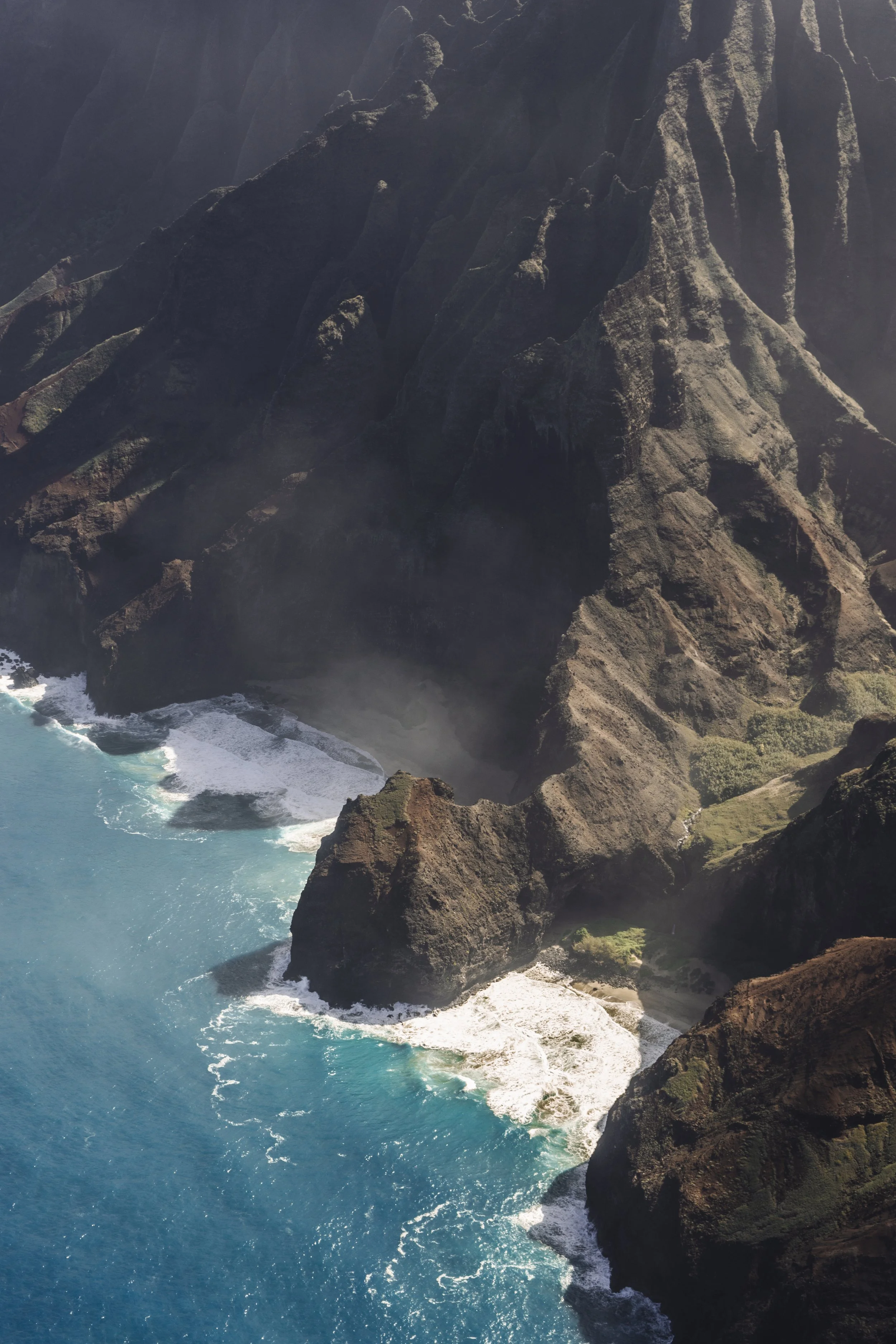 Aerial view of a rocky coastal landscape with steep cliffs, a beach, and turquoise ocean waves crashing against the shore.