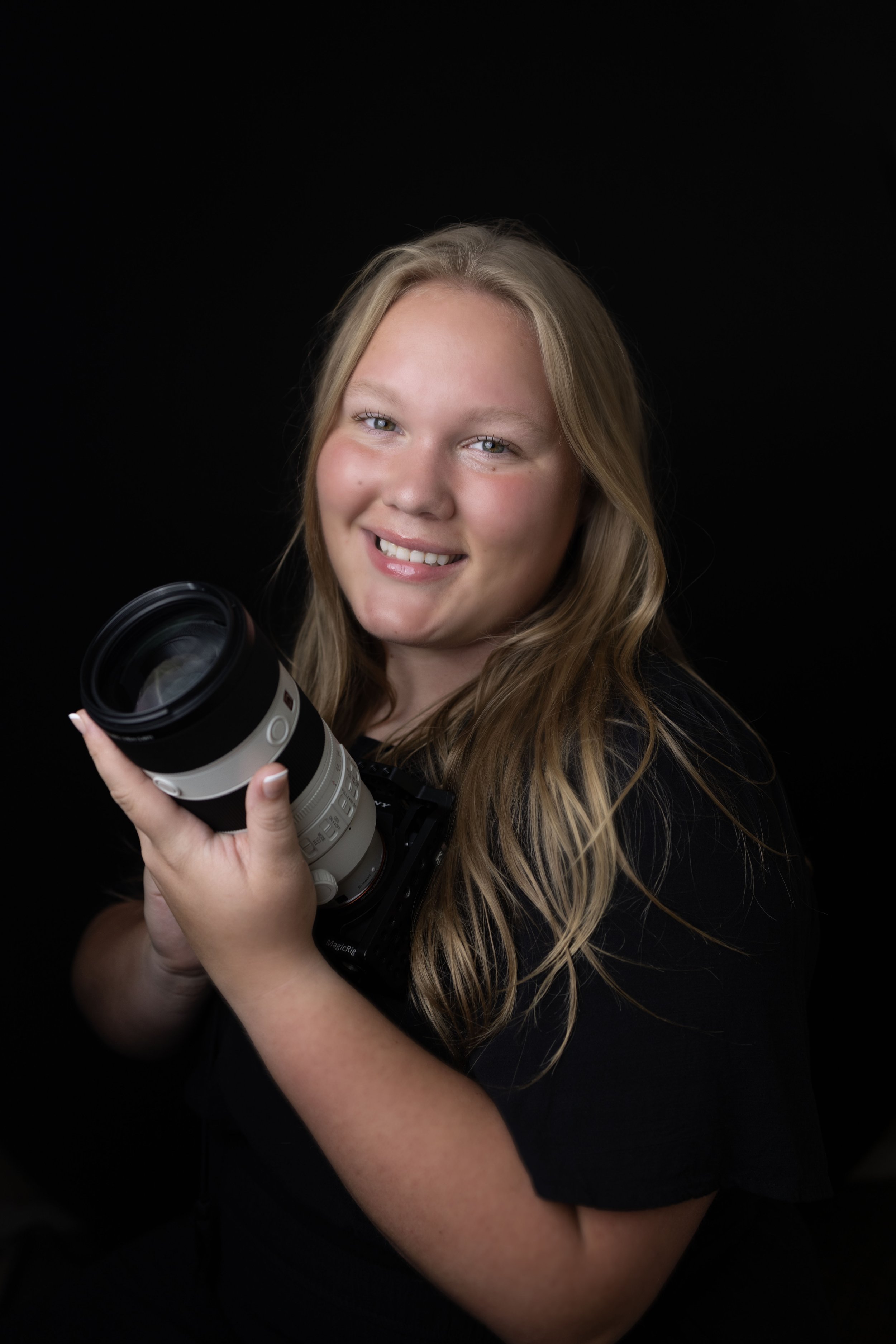A smiling young woman with long blonde hair holding a camera lens against a black background.