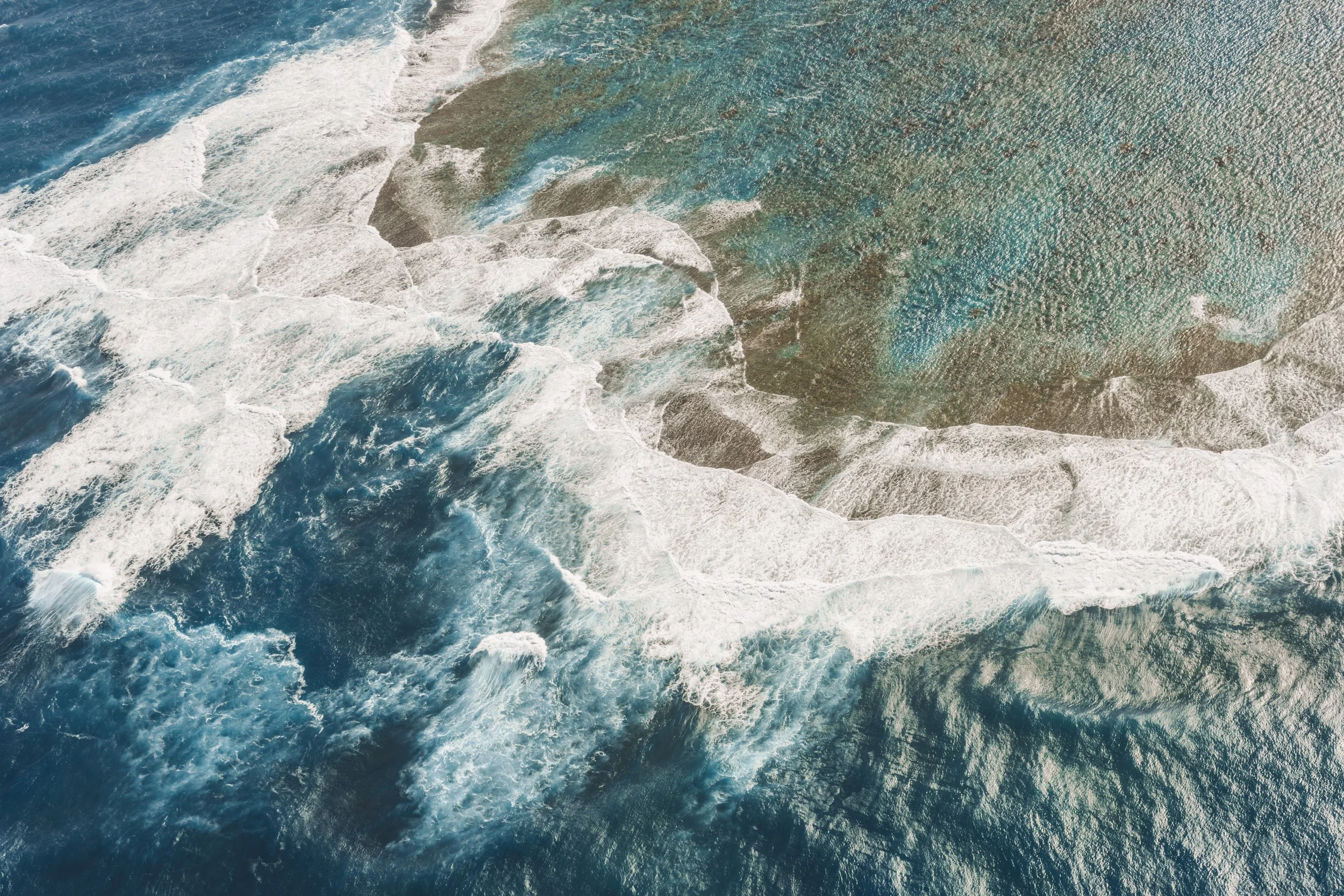 Aerial view of ocean water with white foam and waves crashing over underwater rocks.