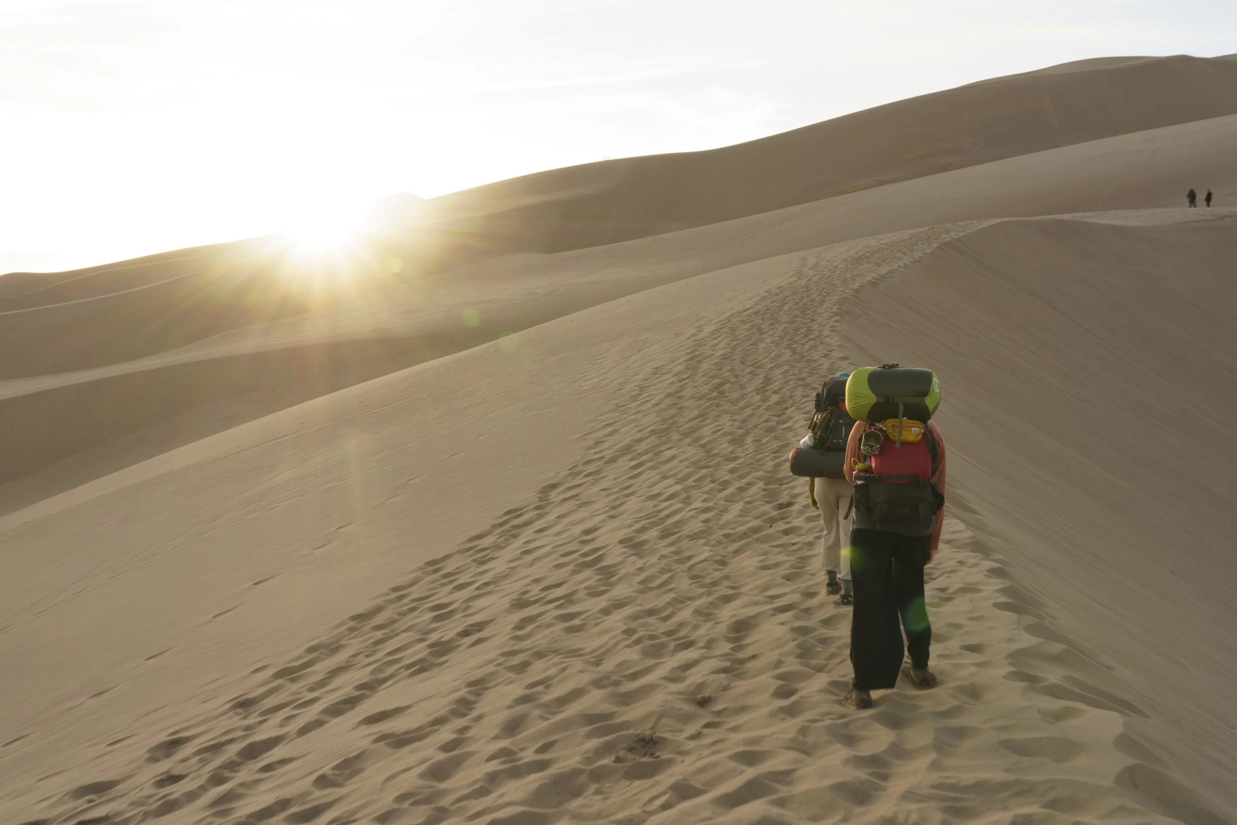 Two hikers with backpacks walking along a sand dune trail in a desert landscape during sunset.