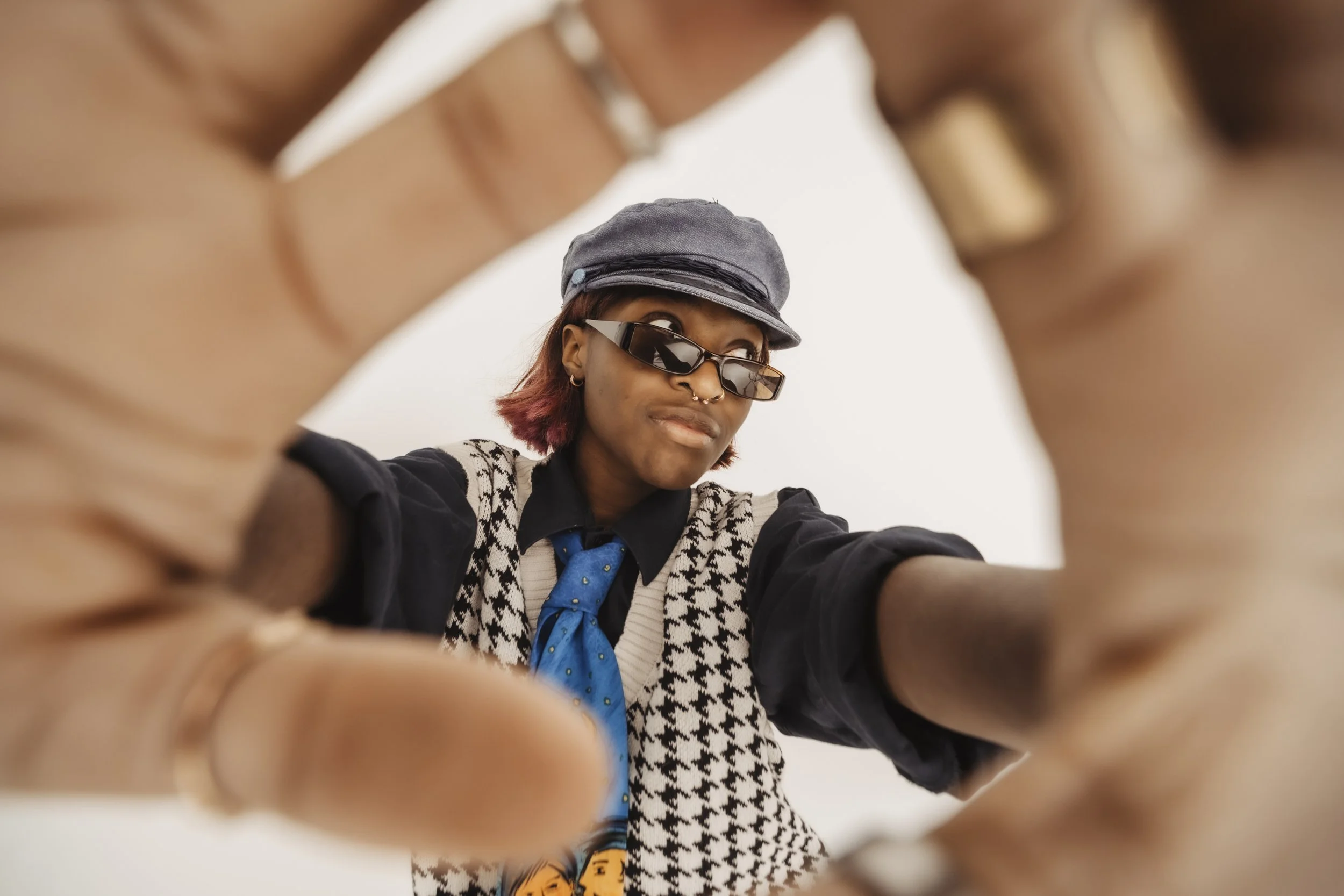 A person with short, dyed red hair wearing sunglasses, a gray cap, a black and white houndstooth vest, a black shirt, and a blue tie, looking down at the camera through their hands, against a white background.