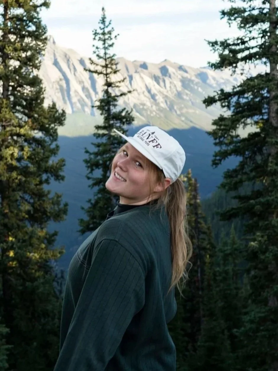 A smiling young woman with long hair in a ponytail, wearing a white cap and a dark jacket, standing outdoors among tall pine trees with a mountain range in the background.