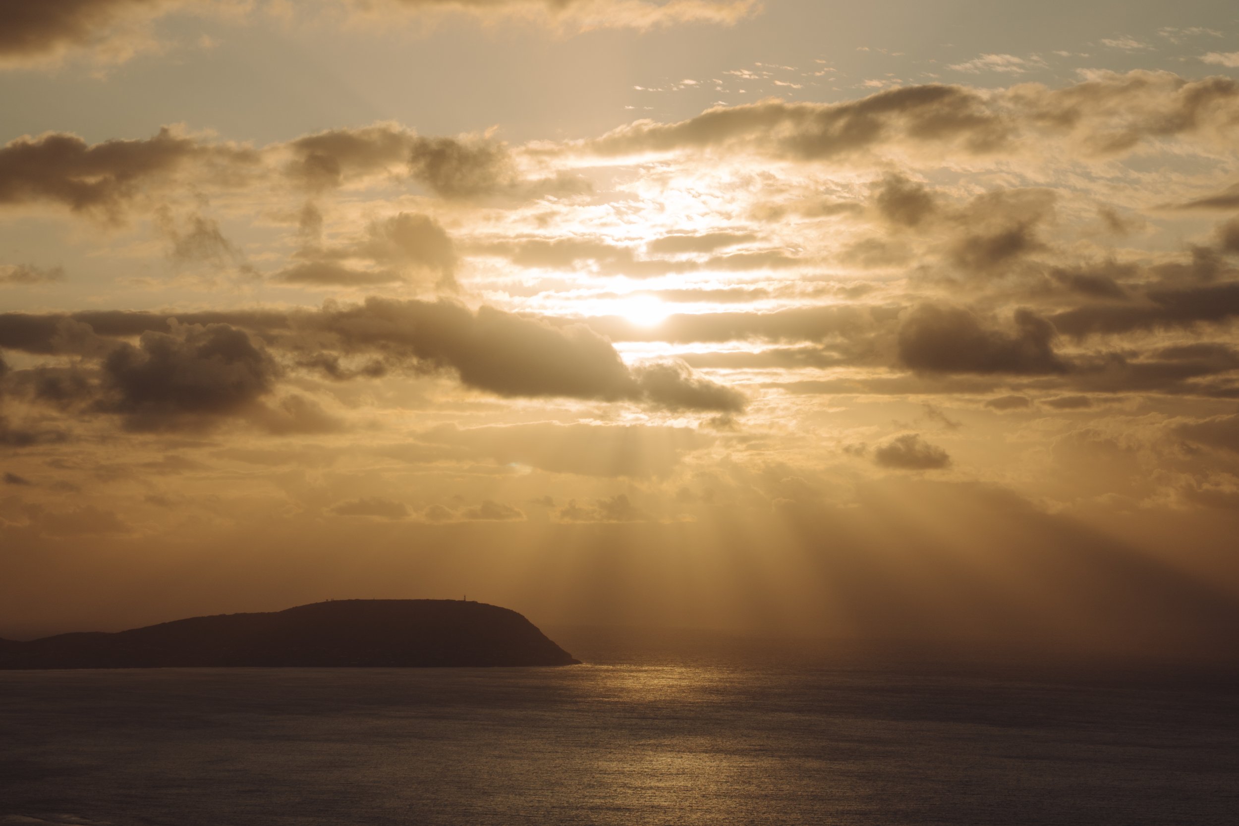 Sunset over the ocean with clouds in the sky and a silhouetted landmass in the distance.