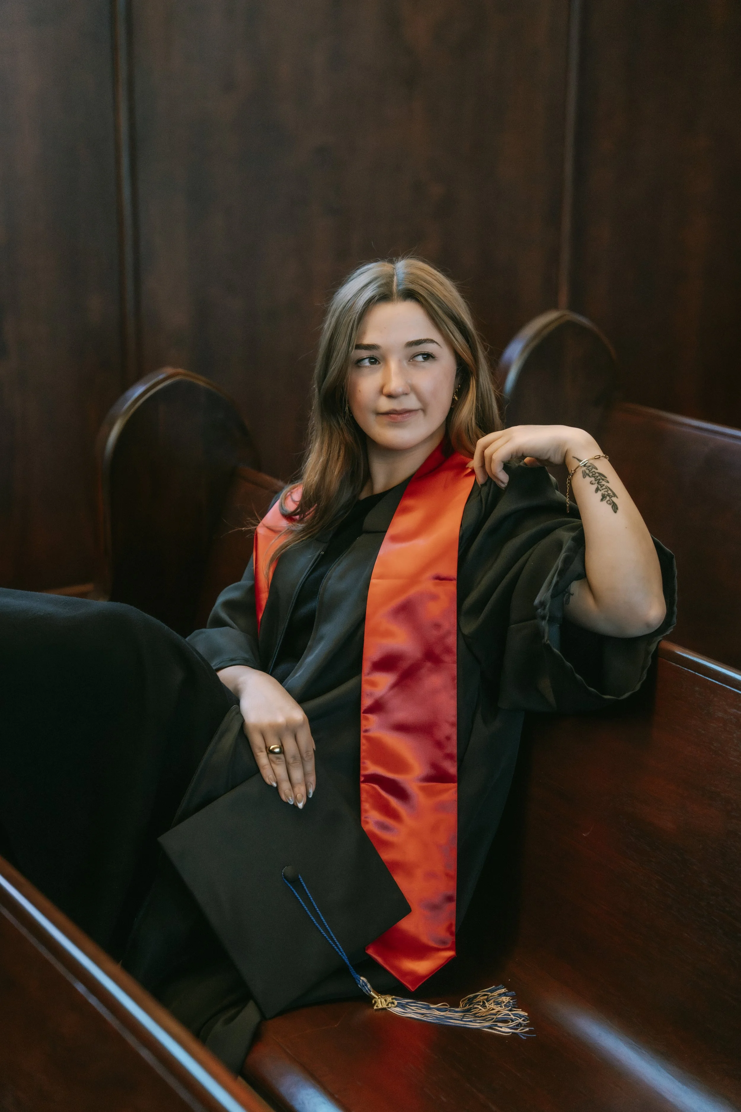 A young woman in a graduation gown with a satin orange sleeve detail, sitting in a wooden pew in a church or auditorium, holding a graduation cap with a blue tassel in her lap.