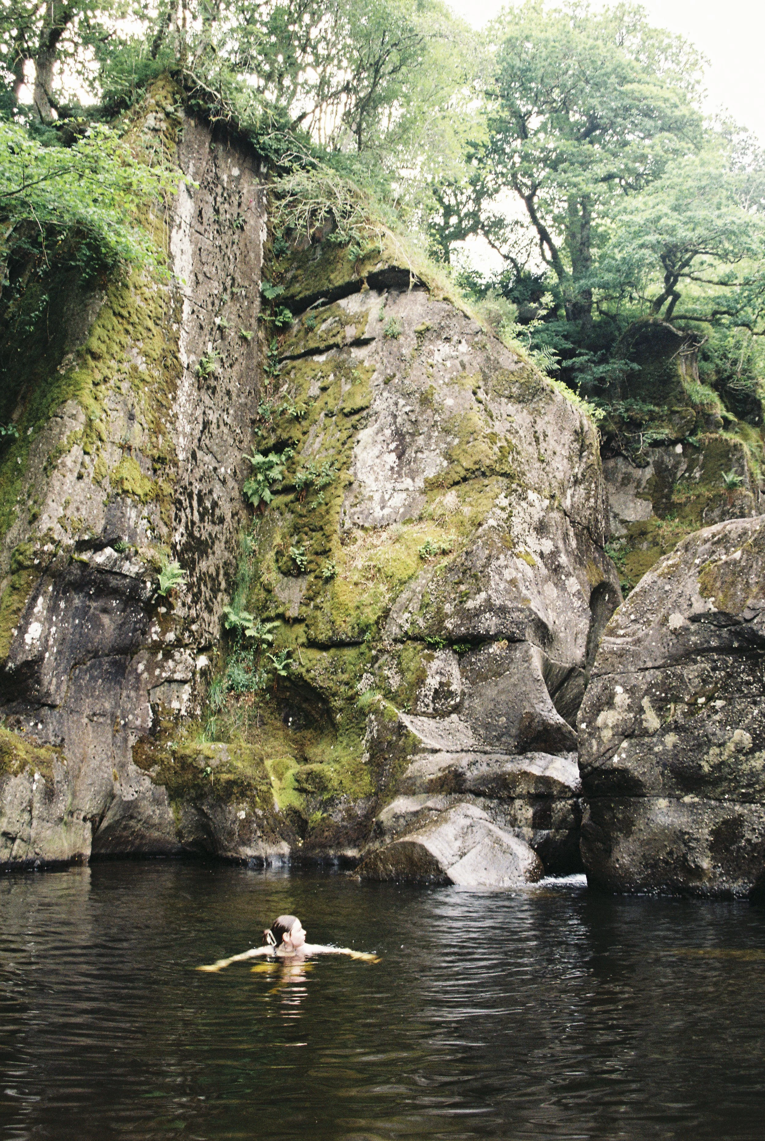 A person swimming in a natural river surrounded by moss-covered rocks and lush green trees.