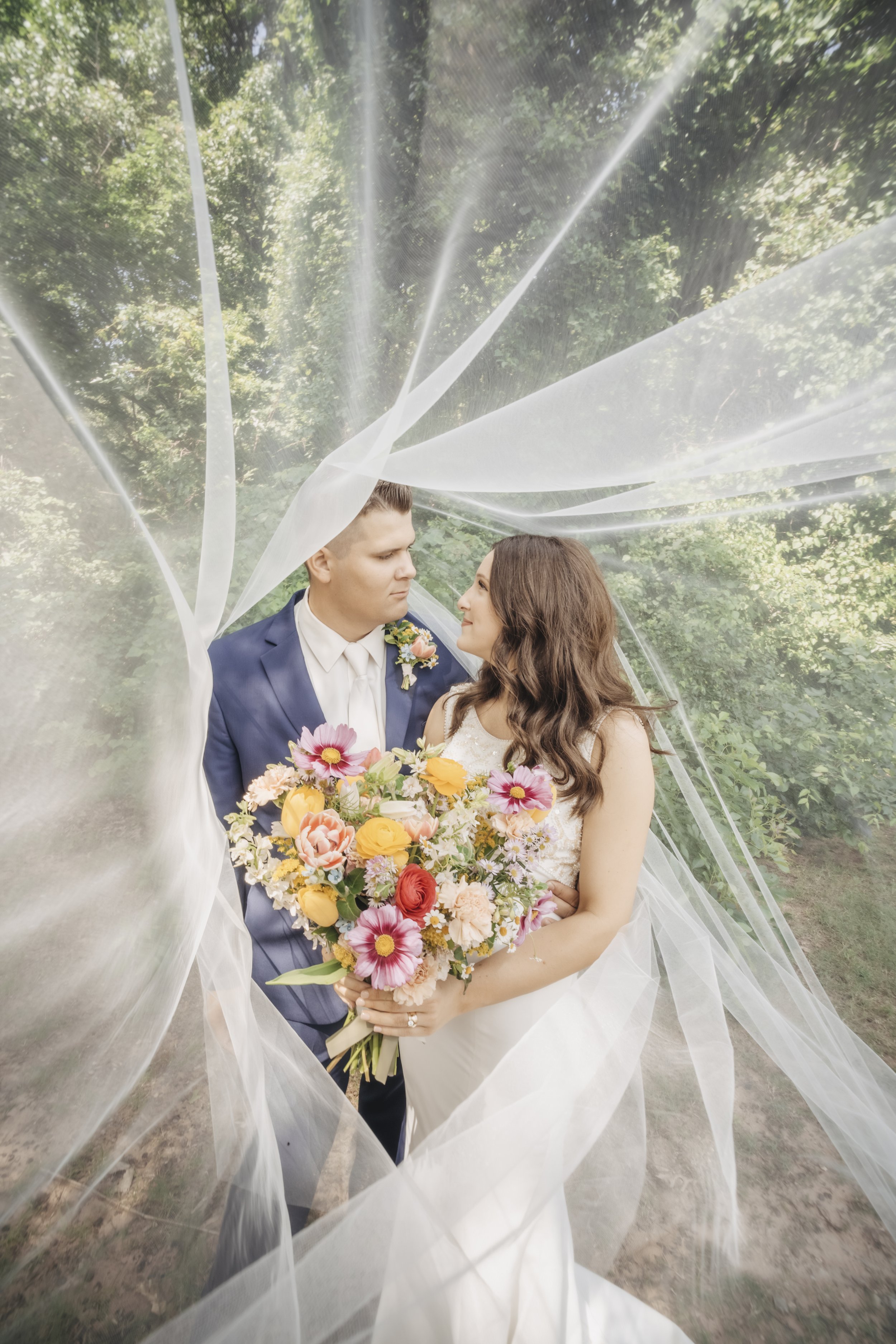 A bride and groom stand together outdoors surrounded by trees, with a veil draped over them. The bride holds a large bouquet of colorful flowers, and they gaze into each other's eyes.