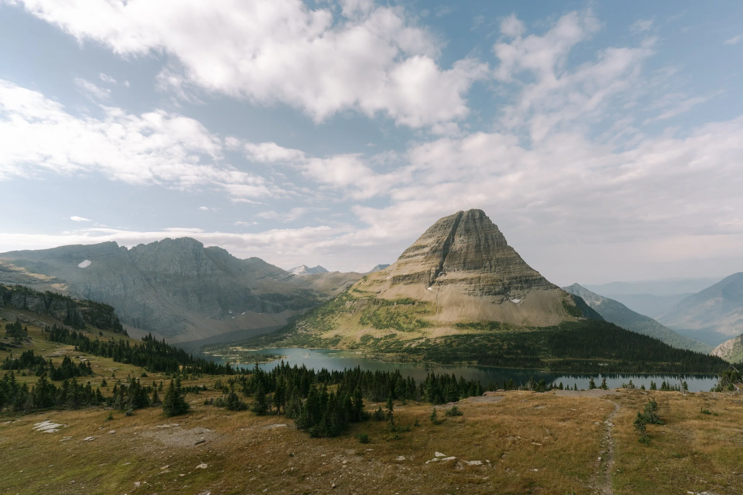 A mountain landscape with a distinctive pyramid-shaped mountain, surrounding hills, a lake, and a partly cloudy sky.