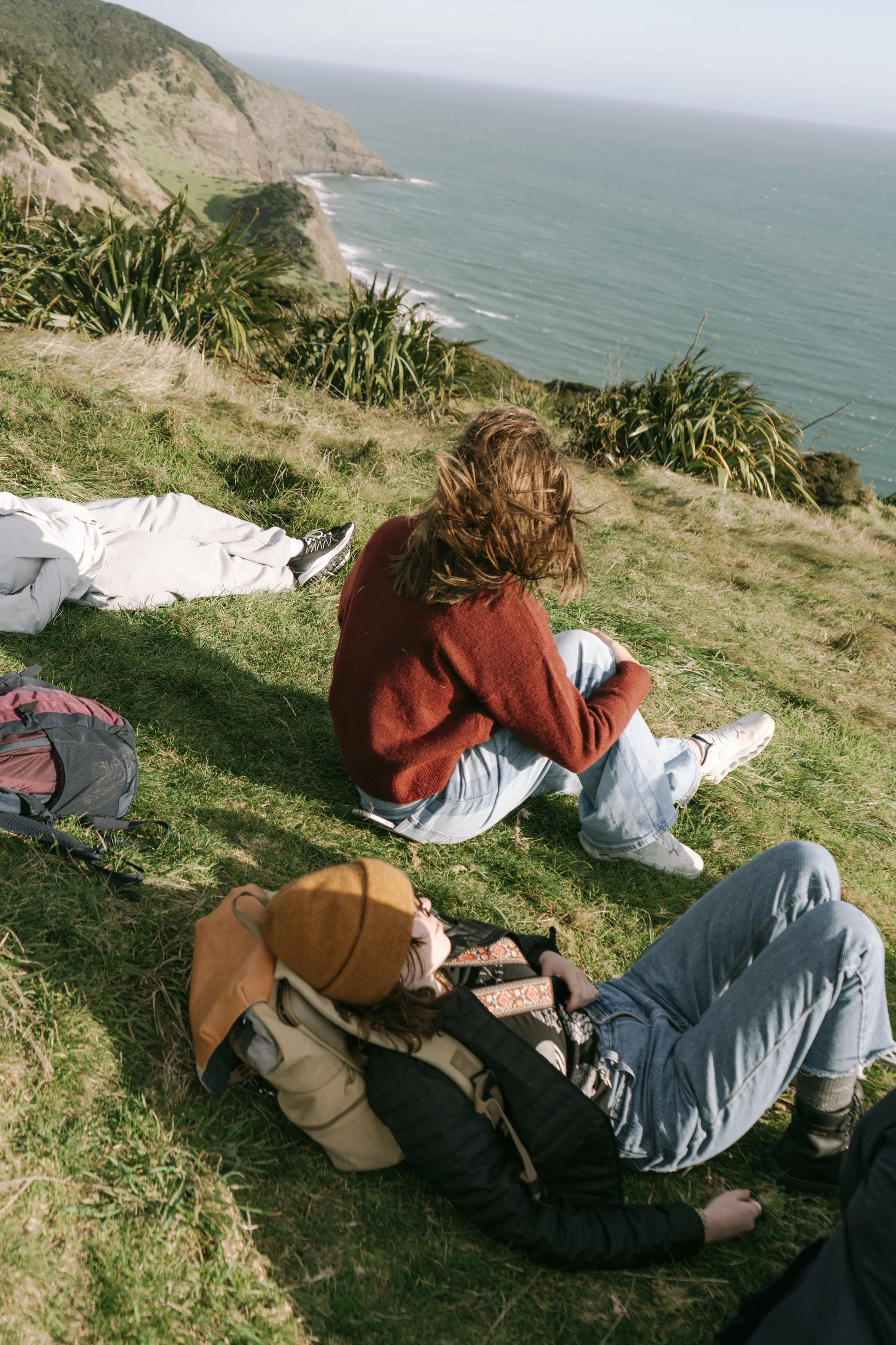 Two people relaxing on a grassy hillside overlooking the ocean with cliffs in the distance, one lying down in a black jacket and brown beanie, the other sitting up in a red sweater.