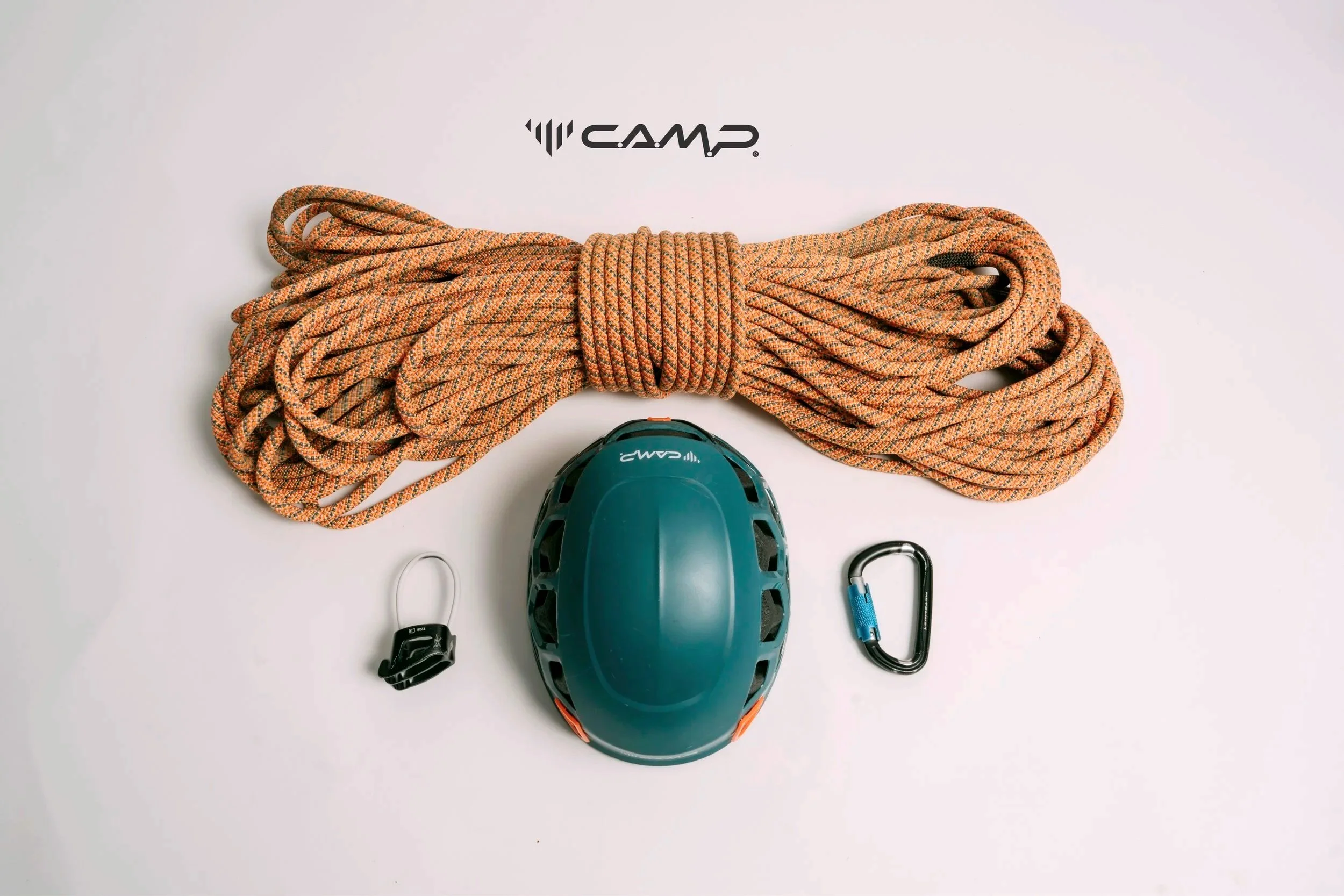 Climbing gear including an orange rope, a green climbing helmet, a black and blue carabiner, and a black adjustable knotter with a small carabiner attached, arranged on a light background with the brand name 'CAMP' visible.