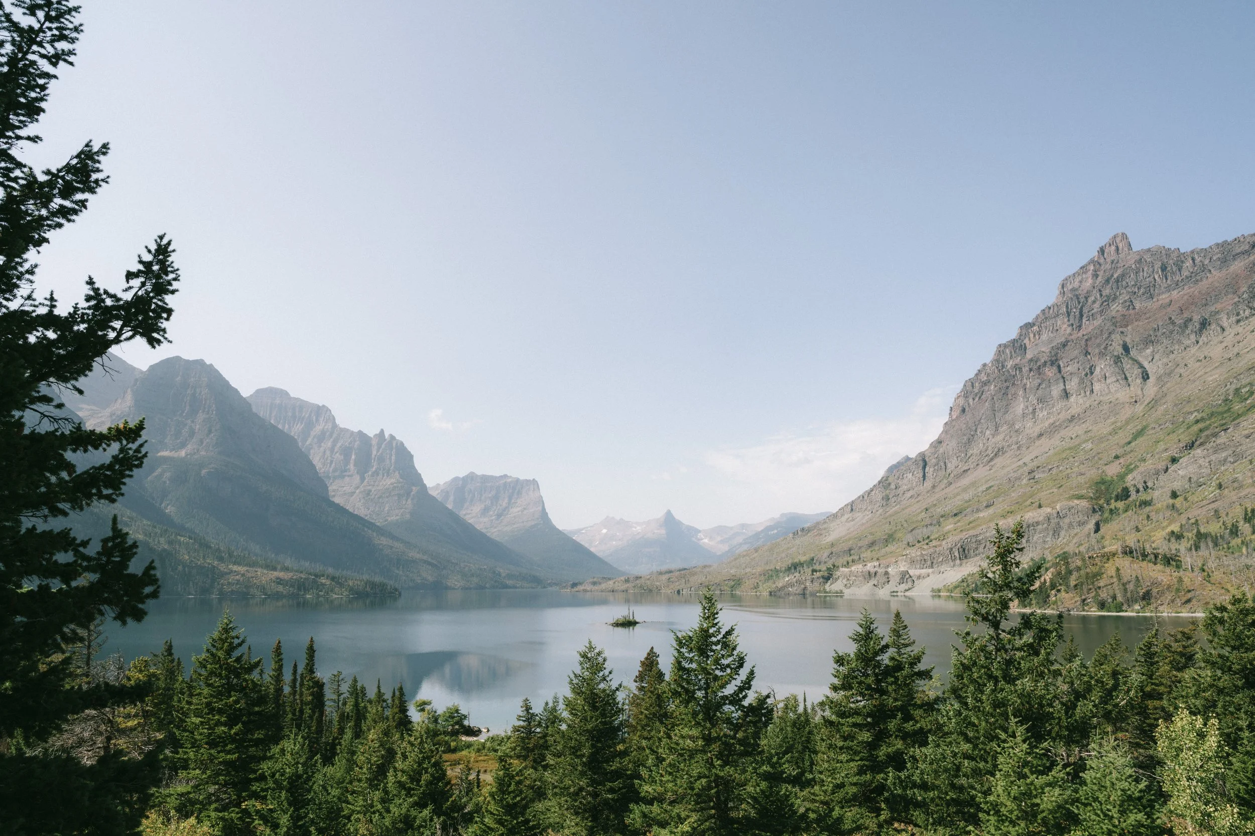 Scenic view of a mountain lake surrounded by evergreen trees, with rugged mountains in the background under a clear blue sky.