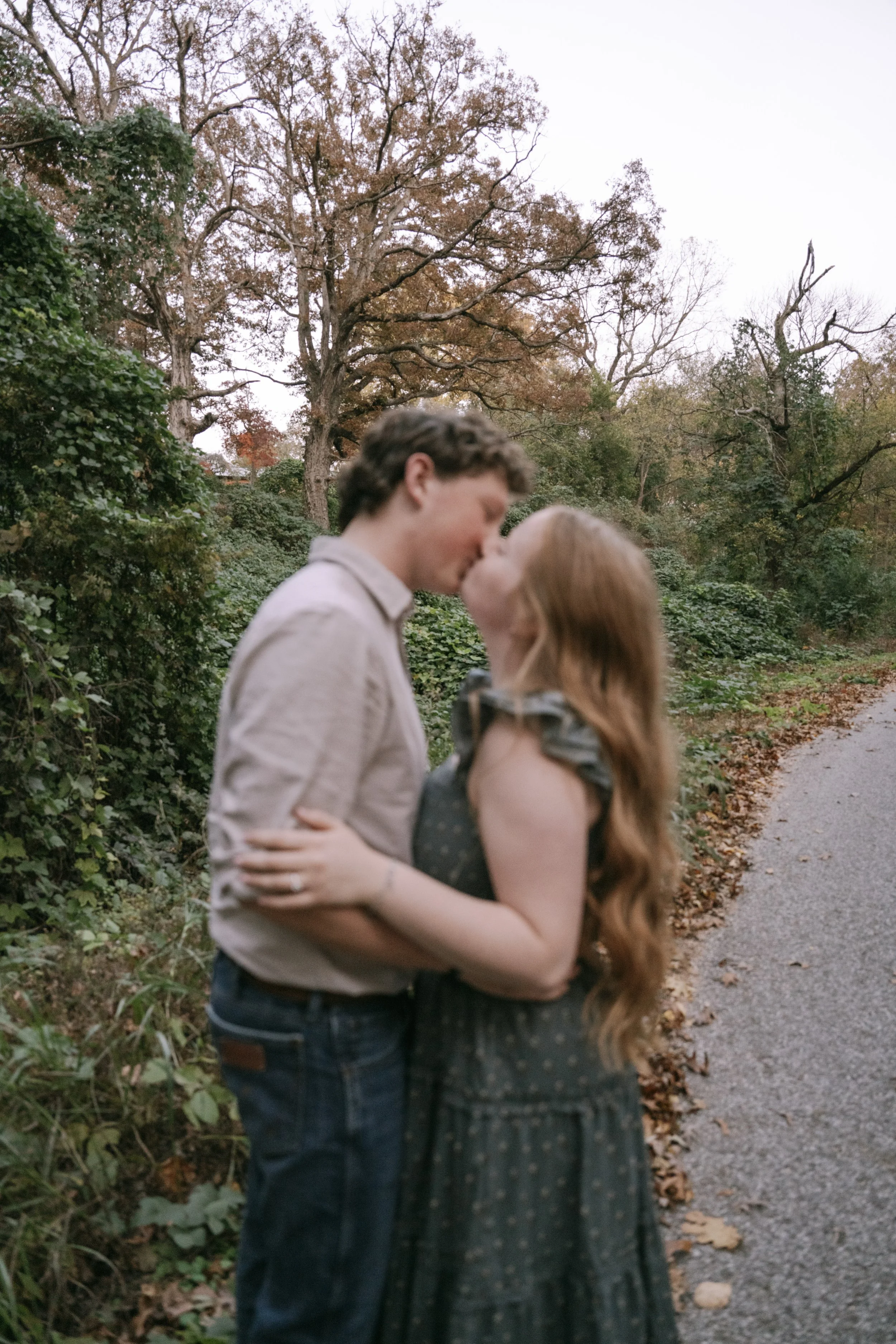 A young couple sharing a kiss outdoors on a gravel path with fall foliage in the background.