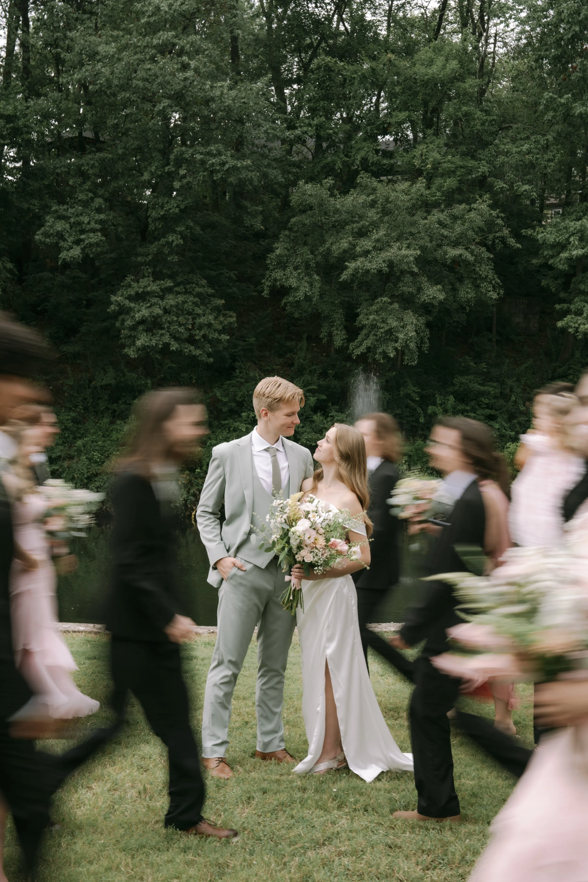 A bride and groom standing together outdoors surrounded by wedding guests holding bouquets, with a lush green wooded background and a small fountain.