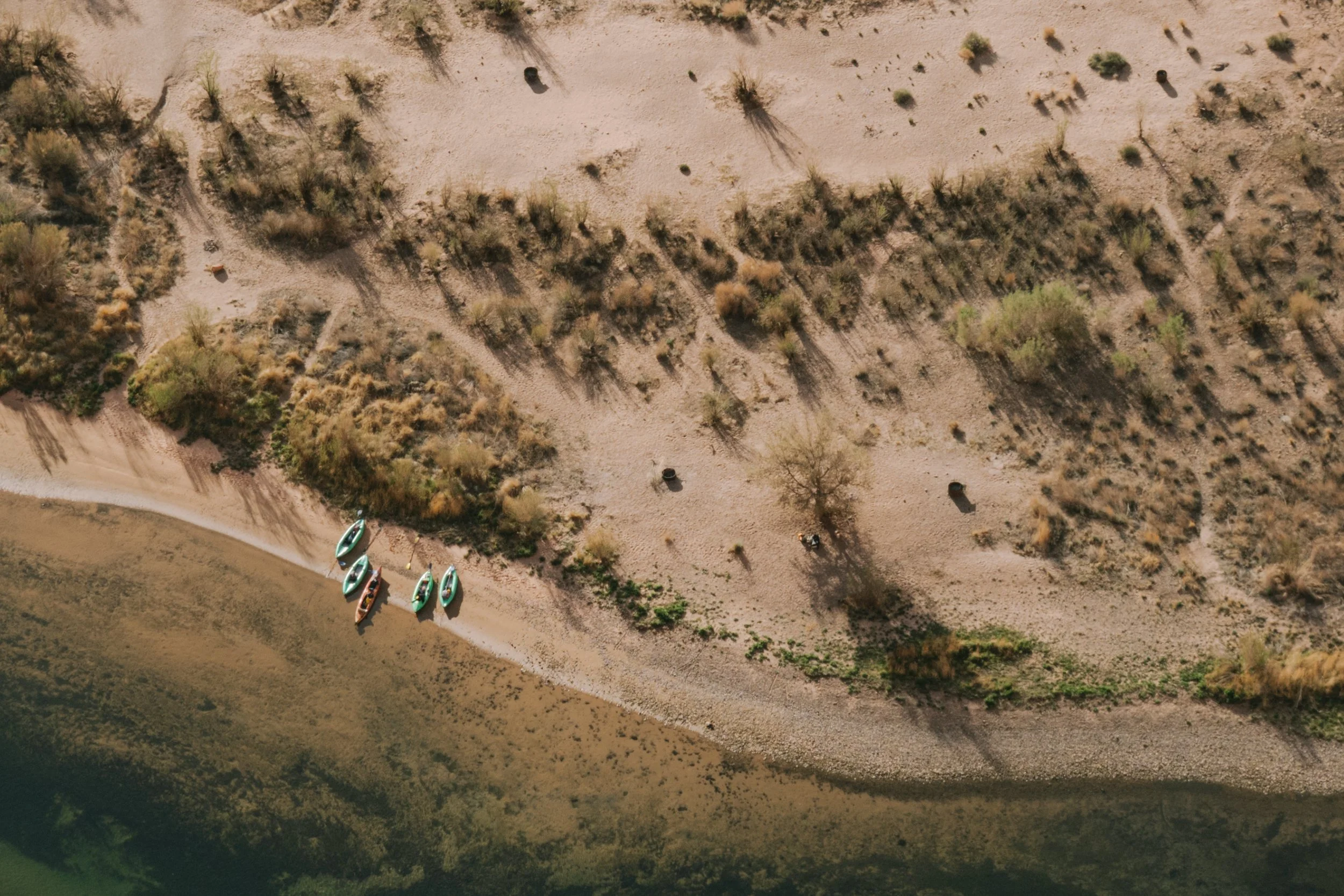 An aerial view of a beach with five kayaks lined up on the sand near the water, sparse vegetation, and shadows cast by trees.