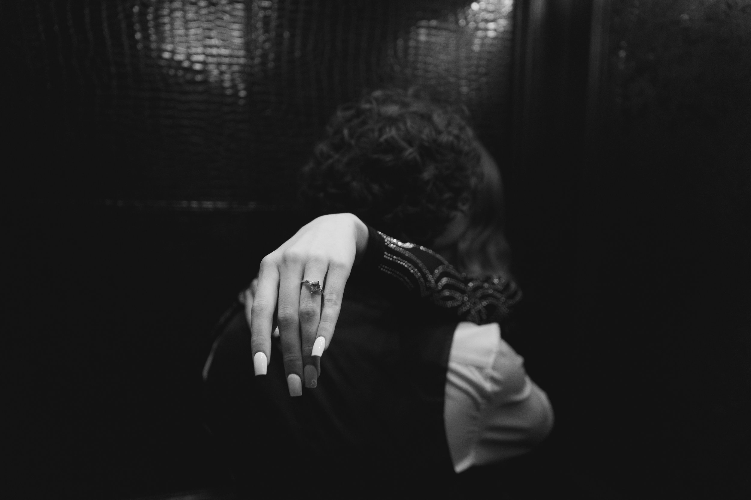 A woman with manicured nails and a ring on her finger is resting her arm on a person's shoulder. The woman has curly hair and is wearing a beaded or embellished top. The background is dark and textured.
