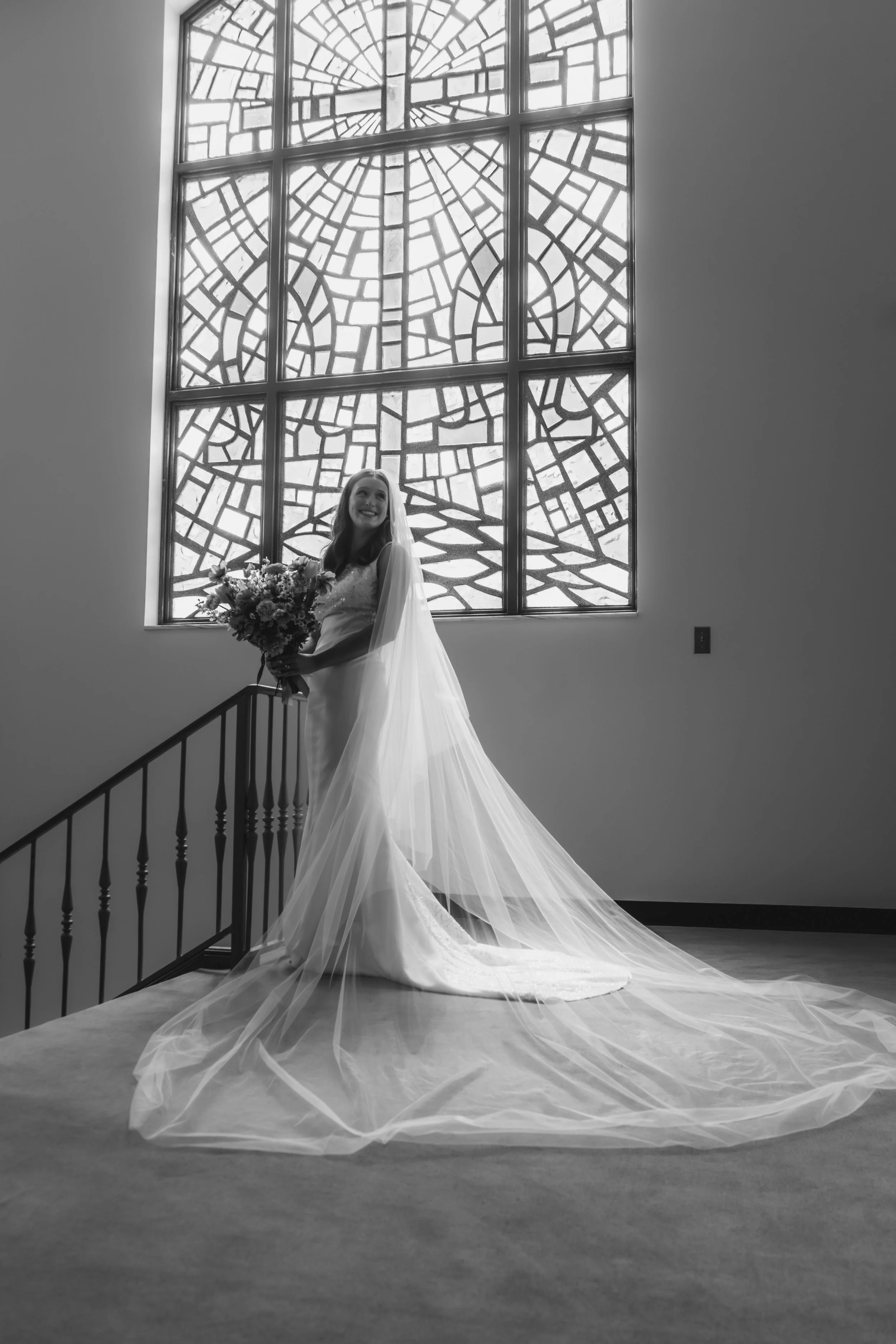 A bride standing indoors on a staircase in front of a large stained glass window, holding a bouquet of flowers, wearing a wedding dress and veil, smiling.