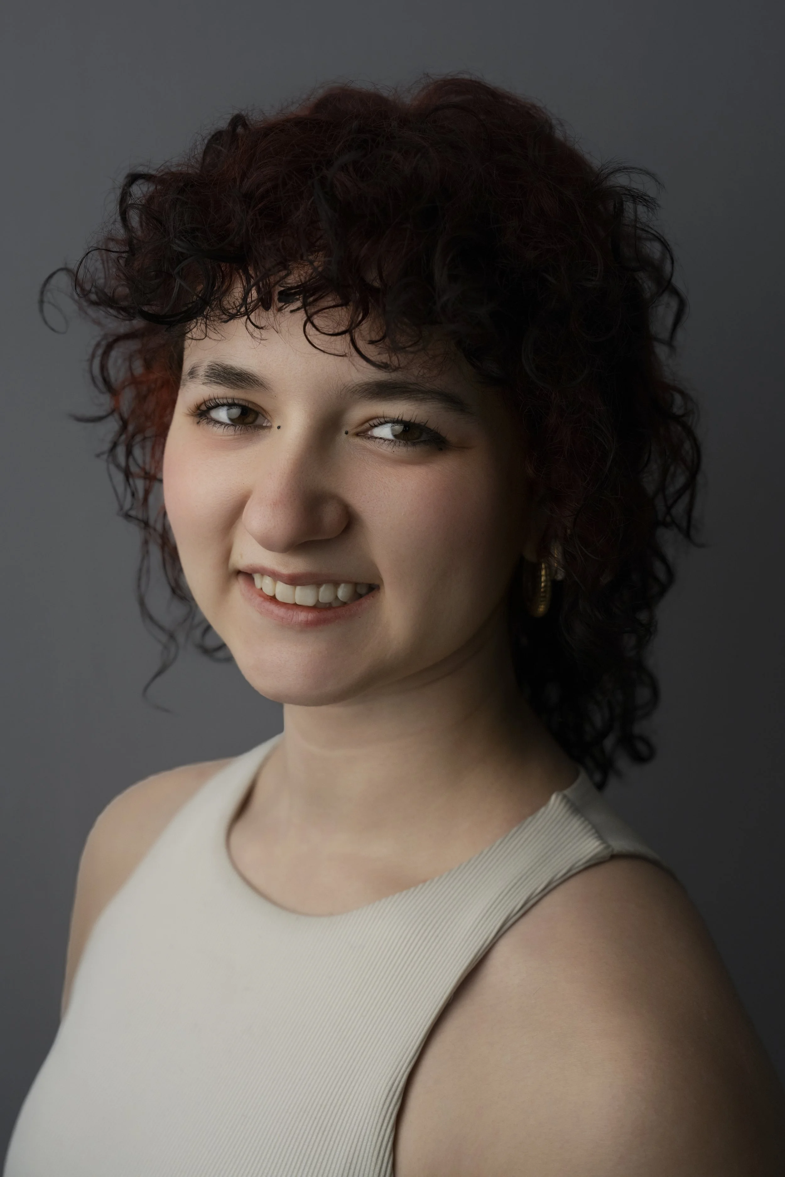 A young woman with short, curly dark hair smiling at the camera, wearing a sleeveless cream top and gold hoop earrings, against a gray background.