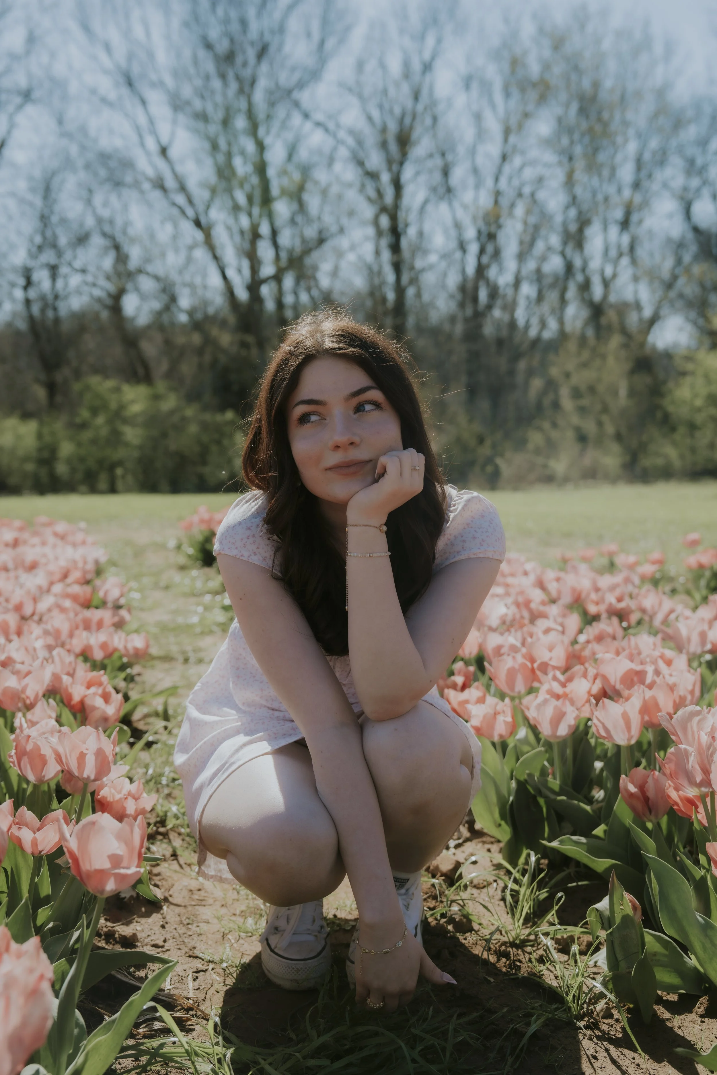 A young woman crouching among pink tulips in a garden during daytime, with trees in the background.